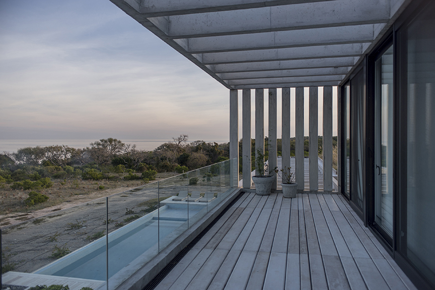 Upper deck under a concrete slab with vertical fins, overlooking a glass-railed lap pool and coastal scrub at dusk