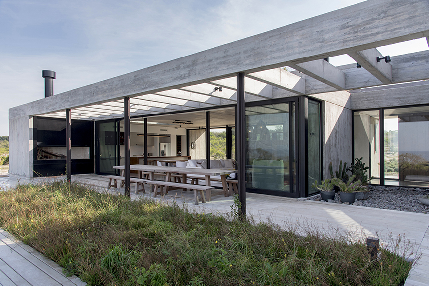 Covered terrace with a long timber dining table beneath a concrete pergola, set behind a foreground of tall grass under pale sky