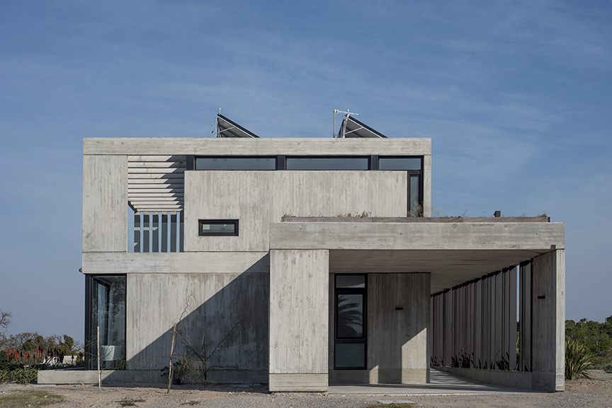 Board-marked concrete facade with a deep carport overhang, rooftop solar heaters and a ribbed screen under a blue streaked sky