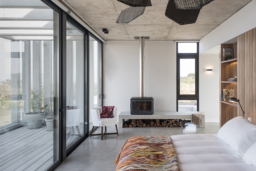 Bedroom with a freestanding steel fireplace above a concrete bench and stacked logs, glass sliding doors opening to a deck in daylight