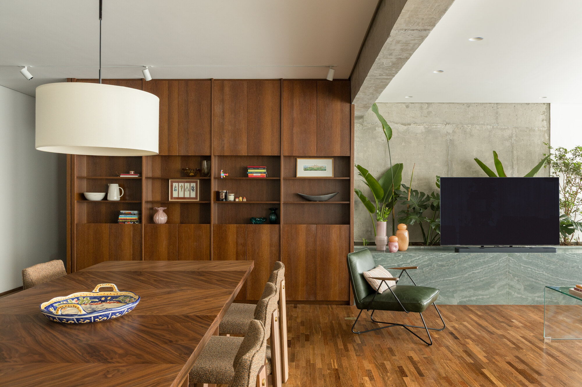 Walnut shelving wall beside a green marble ledge with banana plants, a green leather armchair and a drum pendant over the dining table