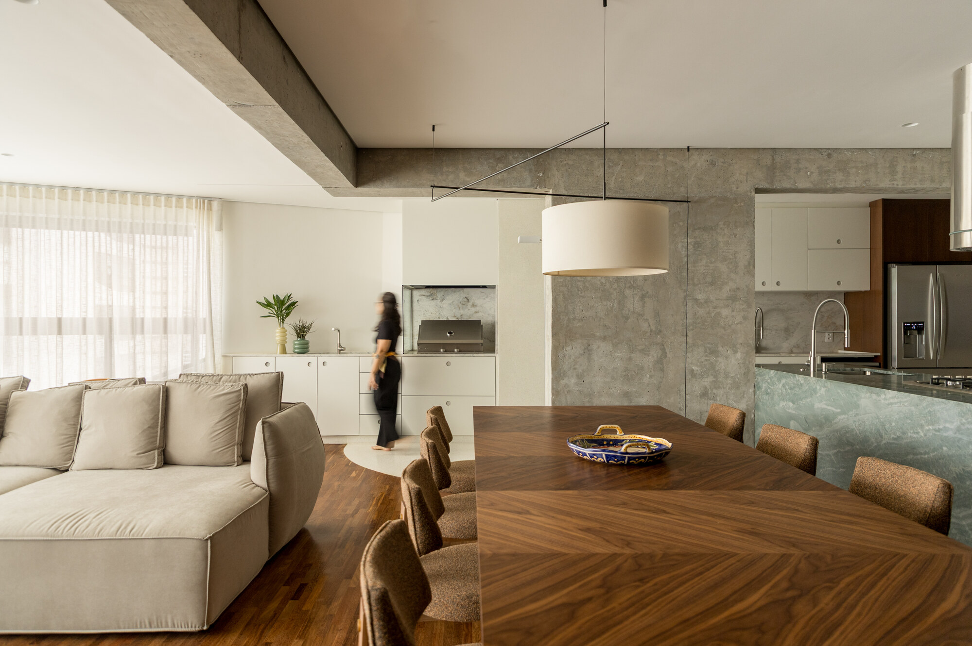 Open living and dining space with a cream sofa, exposed concrete beam and a blurred figure crossing near the built-in grill