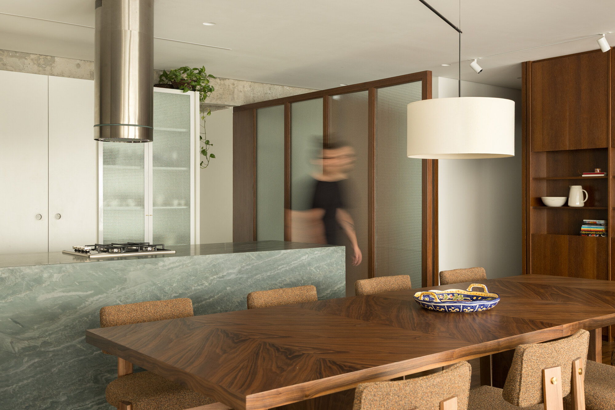 Green marble kitchen island and herringbone walnut table with a blurred figure passing behind ribbed glass partition panels