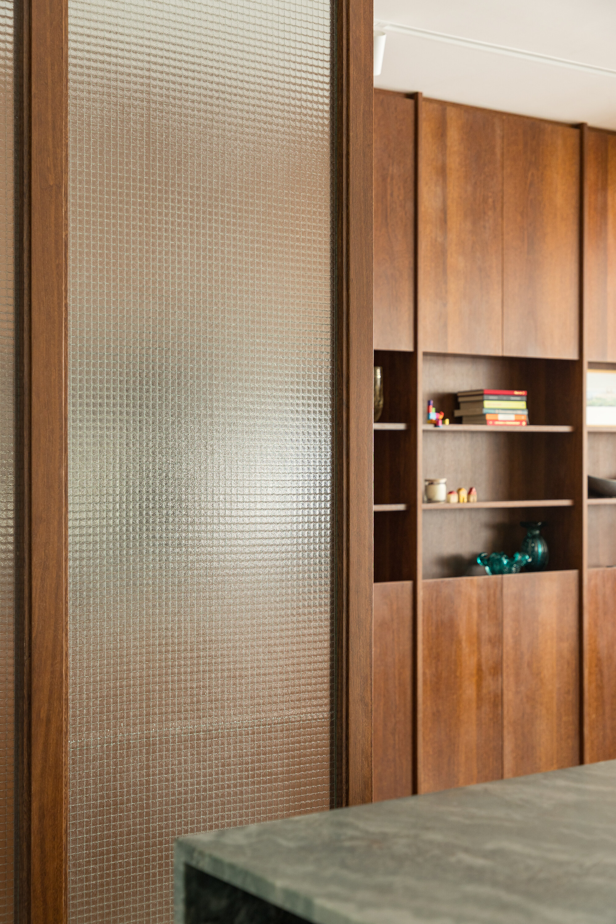 Close-up of the ribbed glass panel in a timber frame beside open shelves holding books and ceramics, with a green stone counter edge