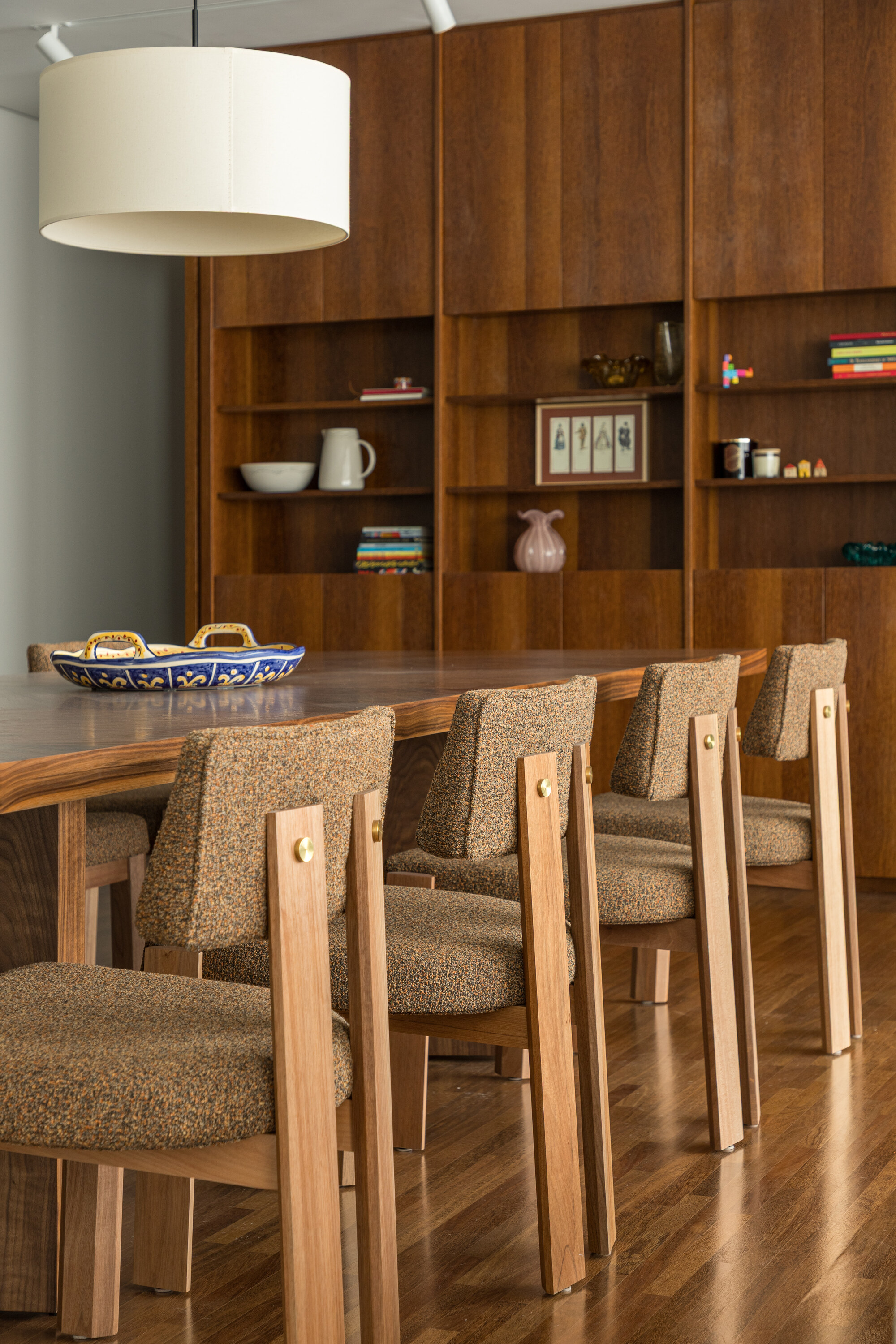 Upholstered timber dining chairs along a wooden table, with a drum pendant lamp and a shelving wall behind