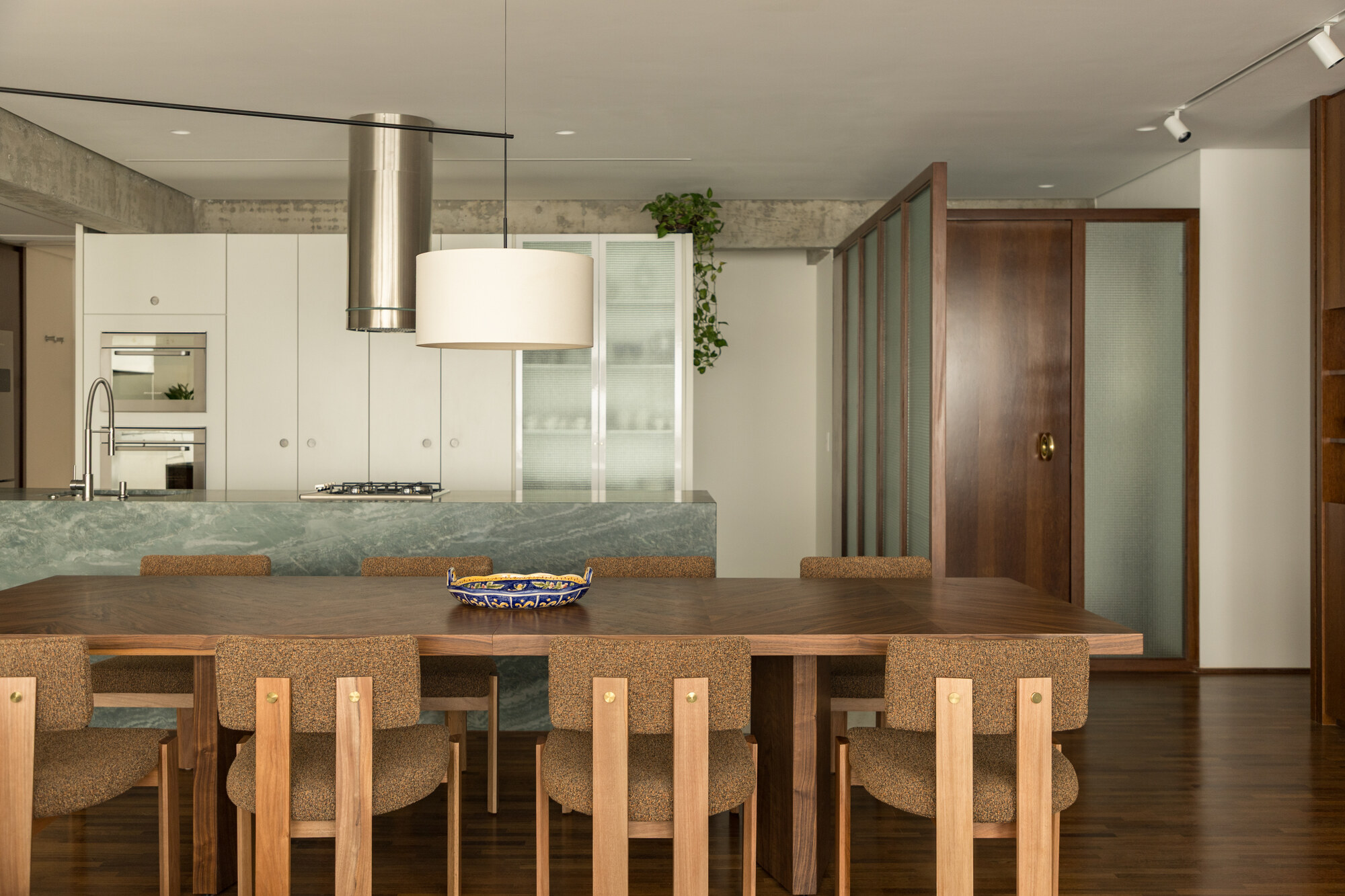 Dining table and chairs facing a green stone kitchen island, with exposed concrete ceiling beams and a timber glazed partition
