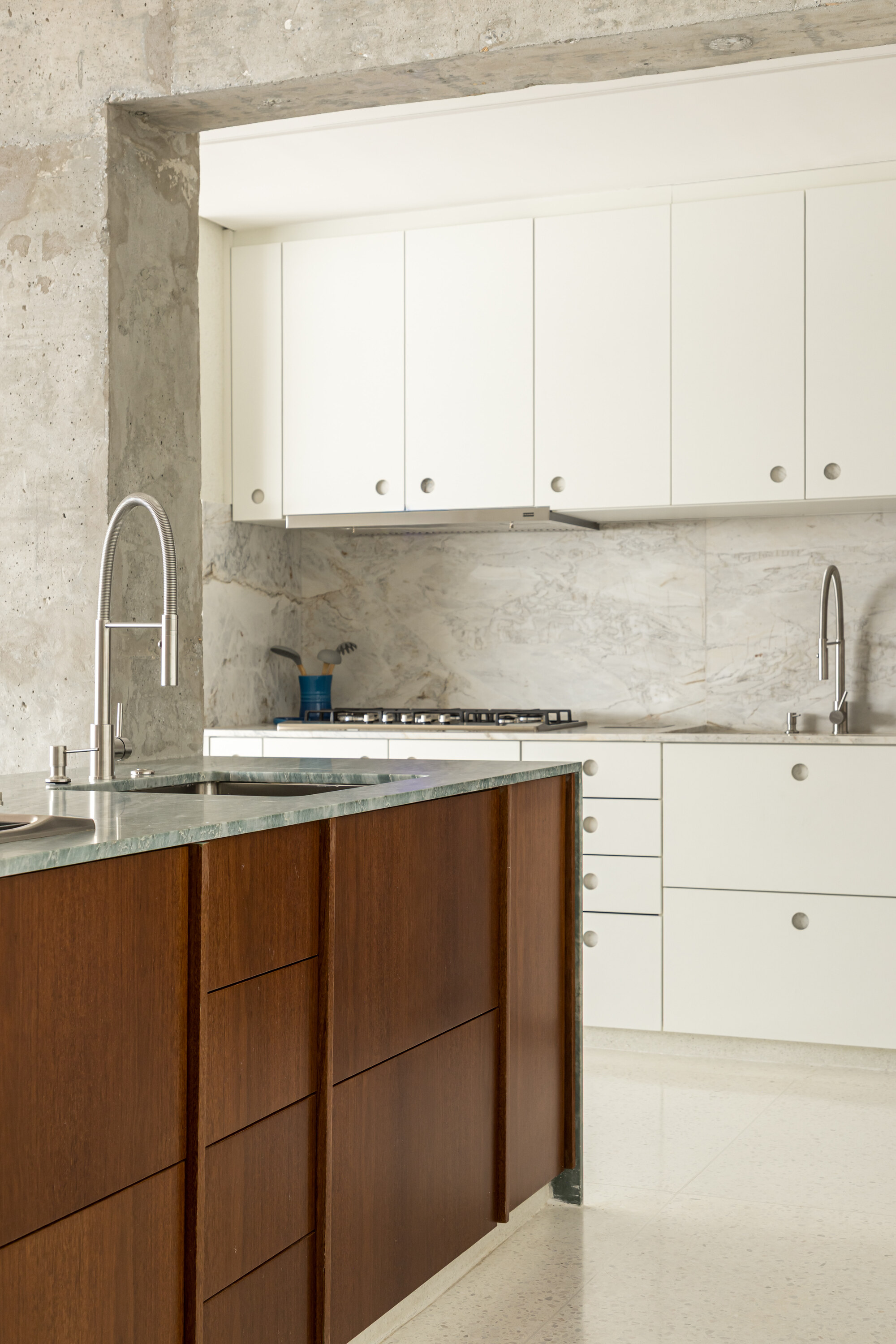 Kitchen corner with white cabinets framed by raw concrete, a marble splashback and a timber island with green stone top