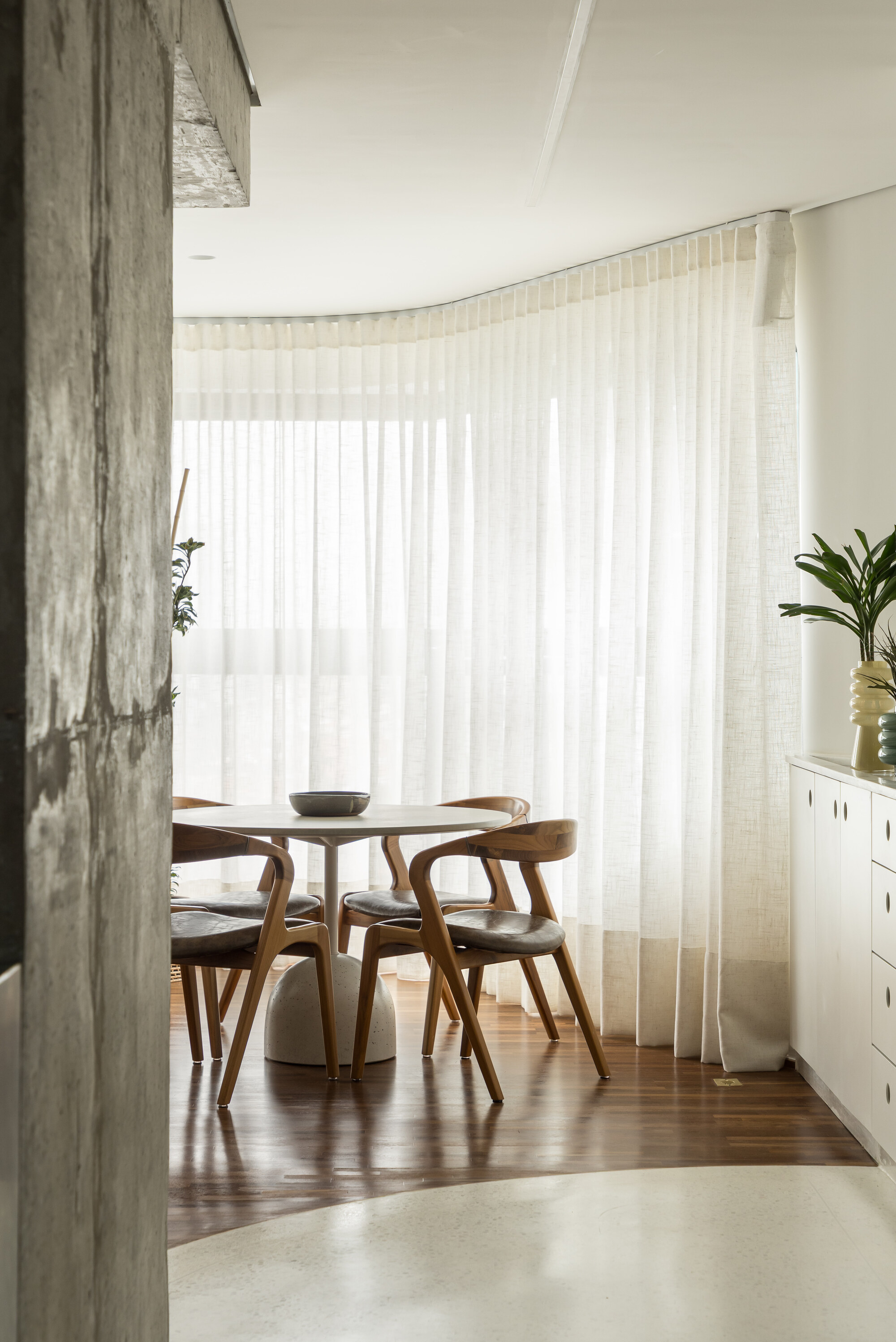 Round dining table with wooden chairs before a curved wall of sheer curtains, beside a rough board-marked concrete column
