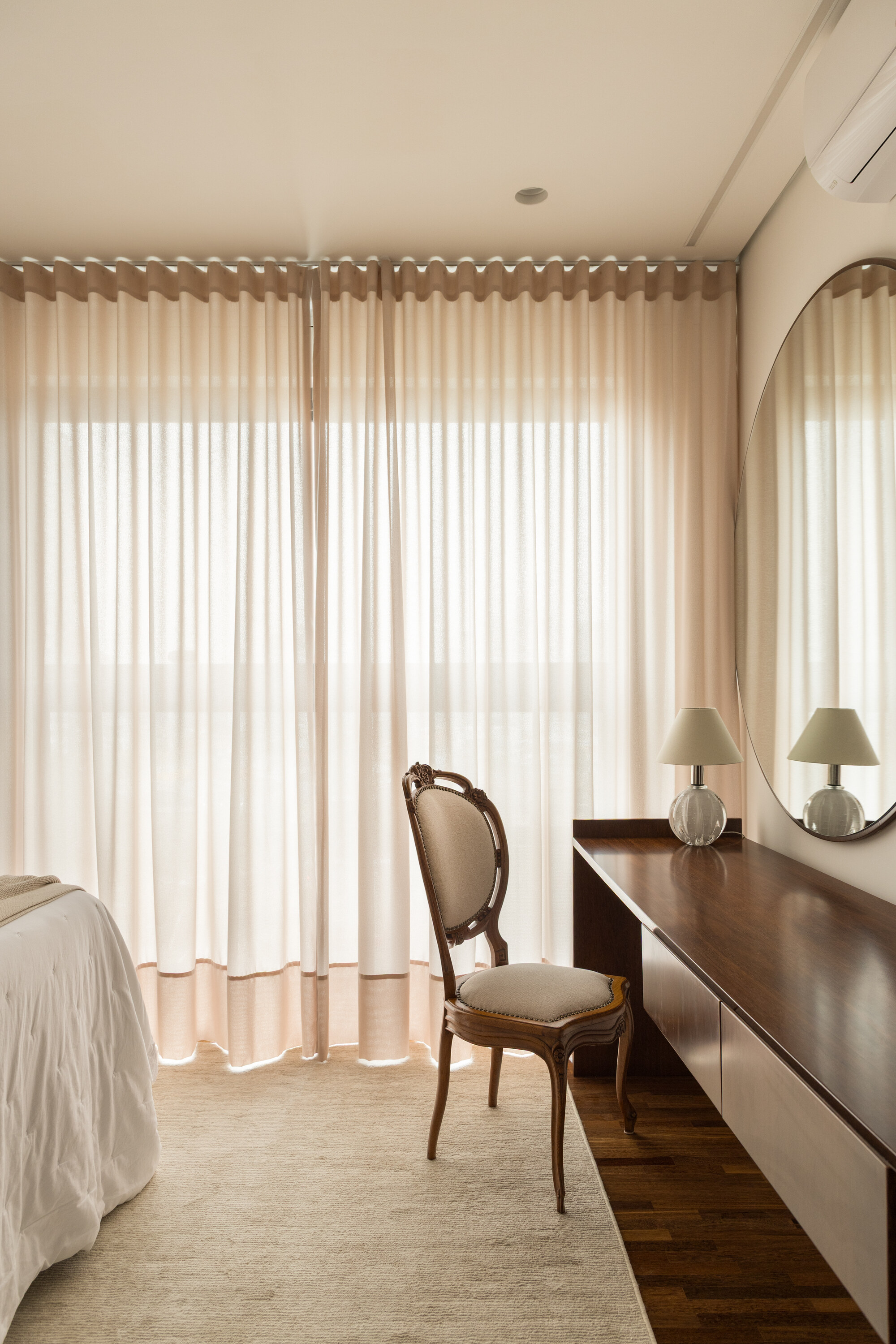 Bedroom corner with a carved wooden chair, long walnut desk and round mirror beside blush pink curtains in diffused light