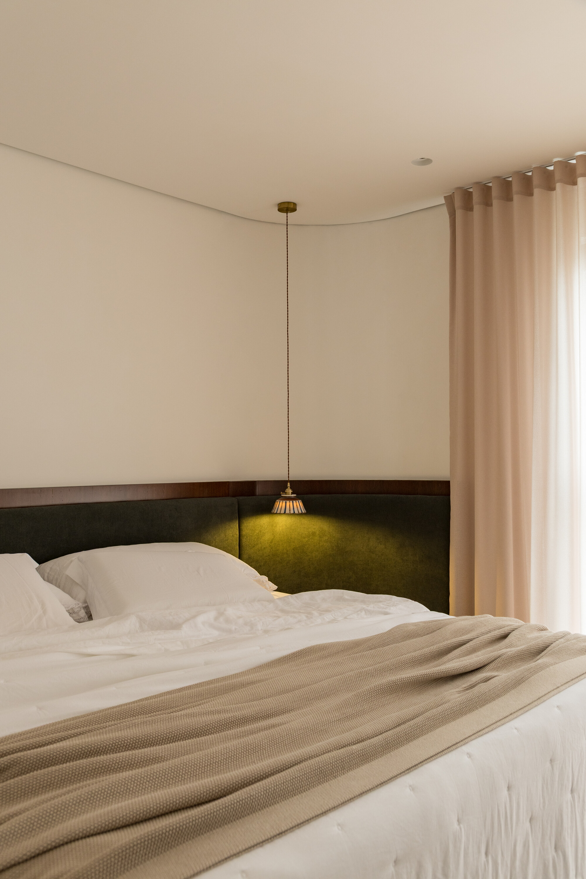 Bed with a dark green upholstered headboard curving along the wall, lit by a brass pendant lamp beside a pink curtain