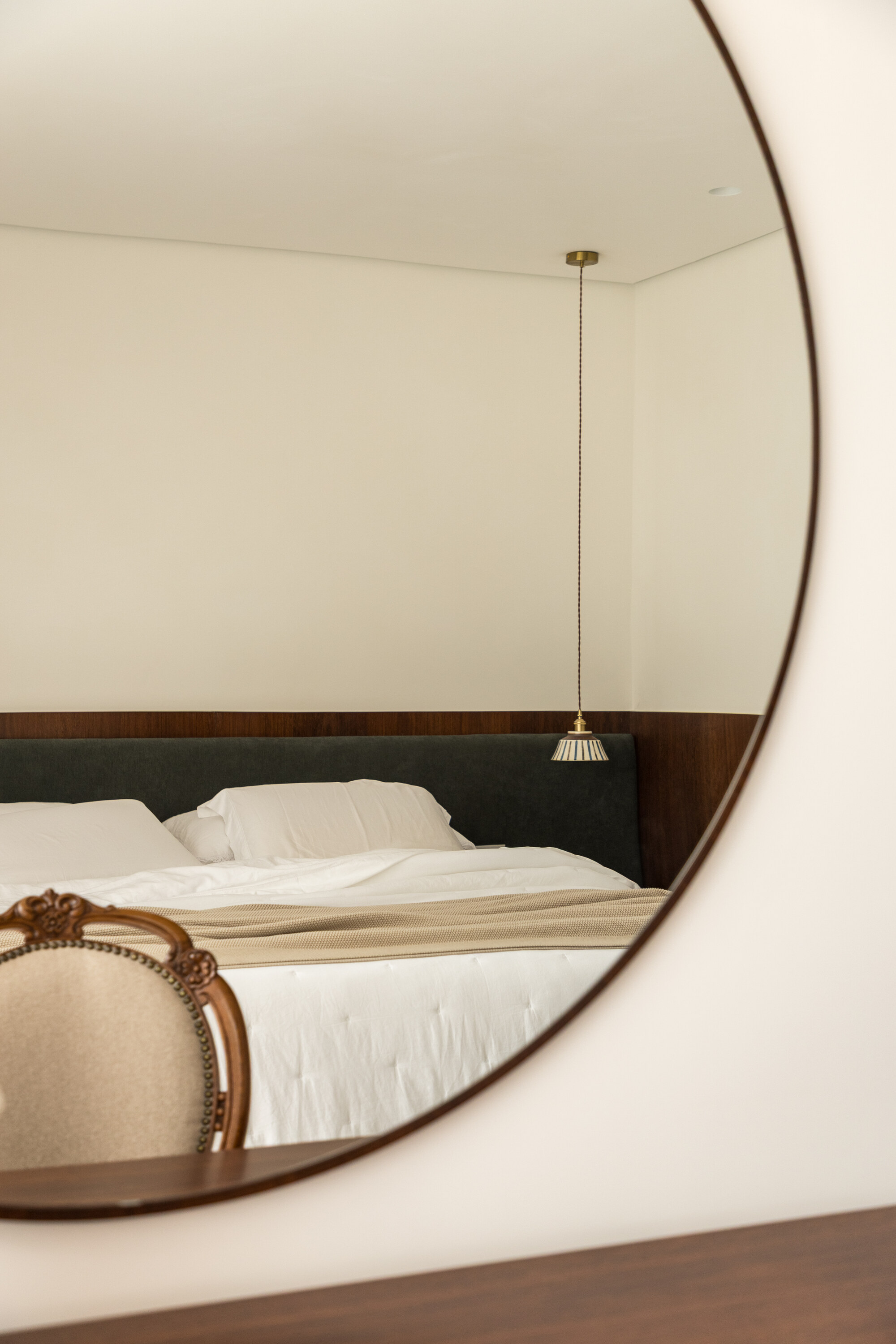 Round mirror reflecting the bed, green headboard and brass pendant lamp, with the back of a carved chair in the foreground