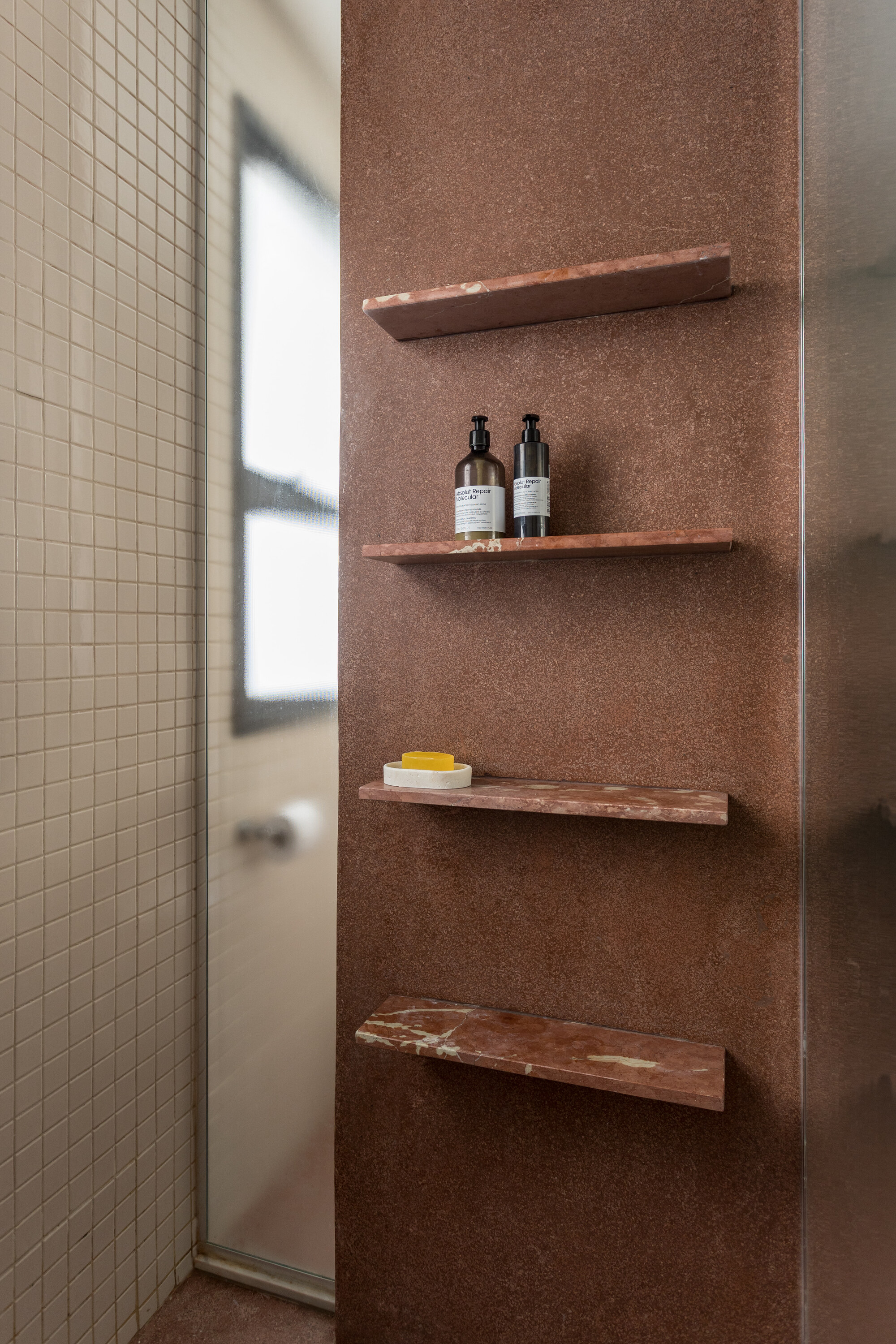 Four red marble shelves holding bottles and a soap dish on a rust-colored shower wall beside small beige tiles