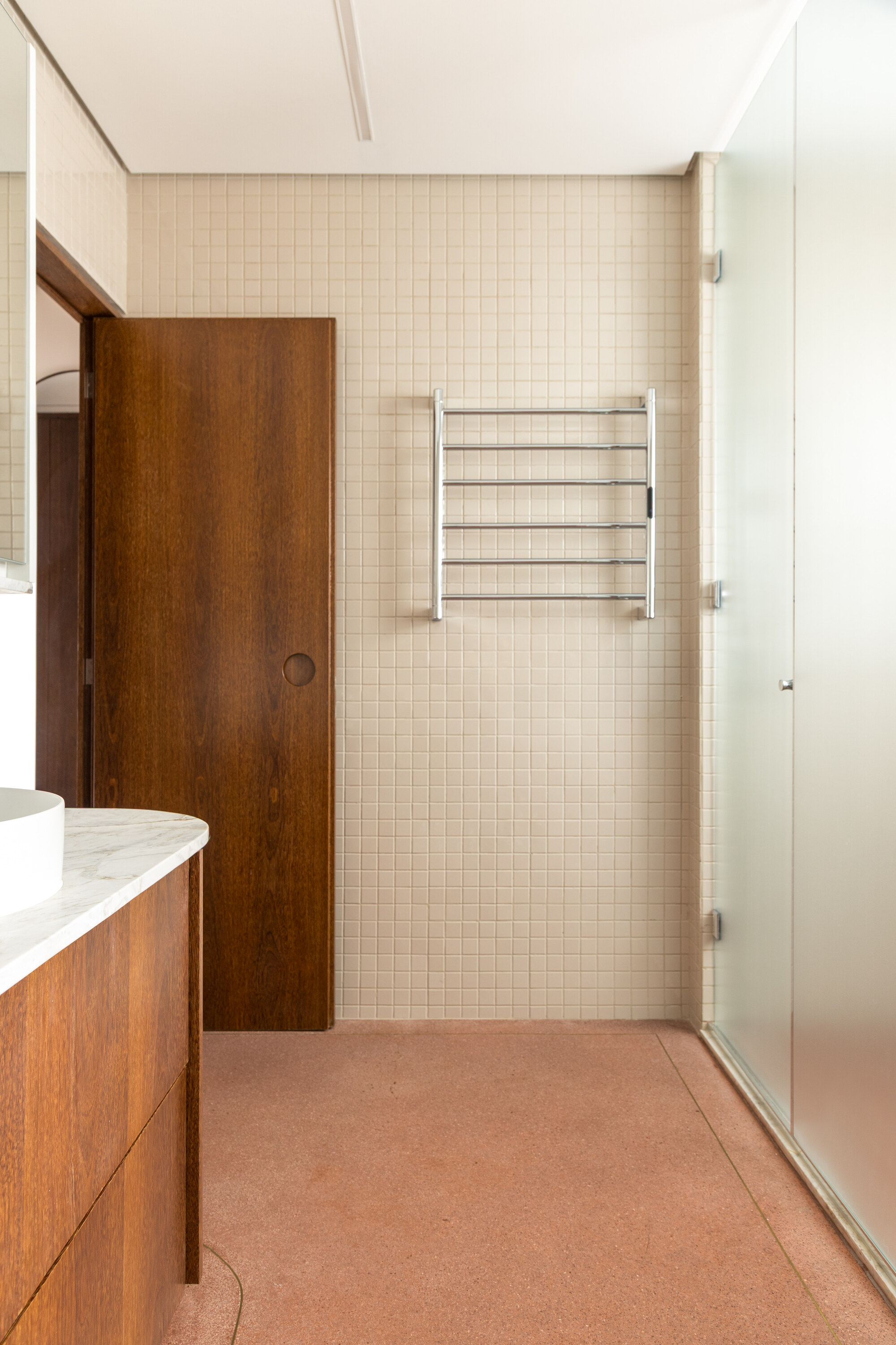 Bathroom wall of small beige tiles with a chrome towel rail, a timber pivot door and frosted glass shower panel