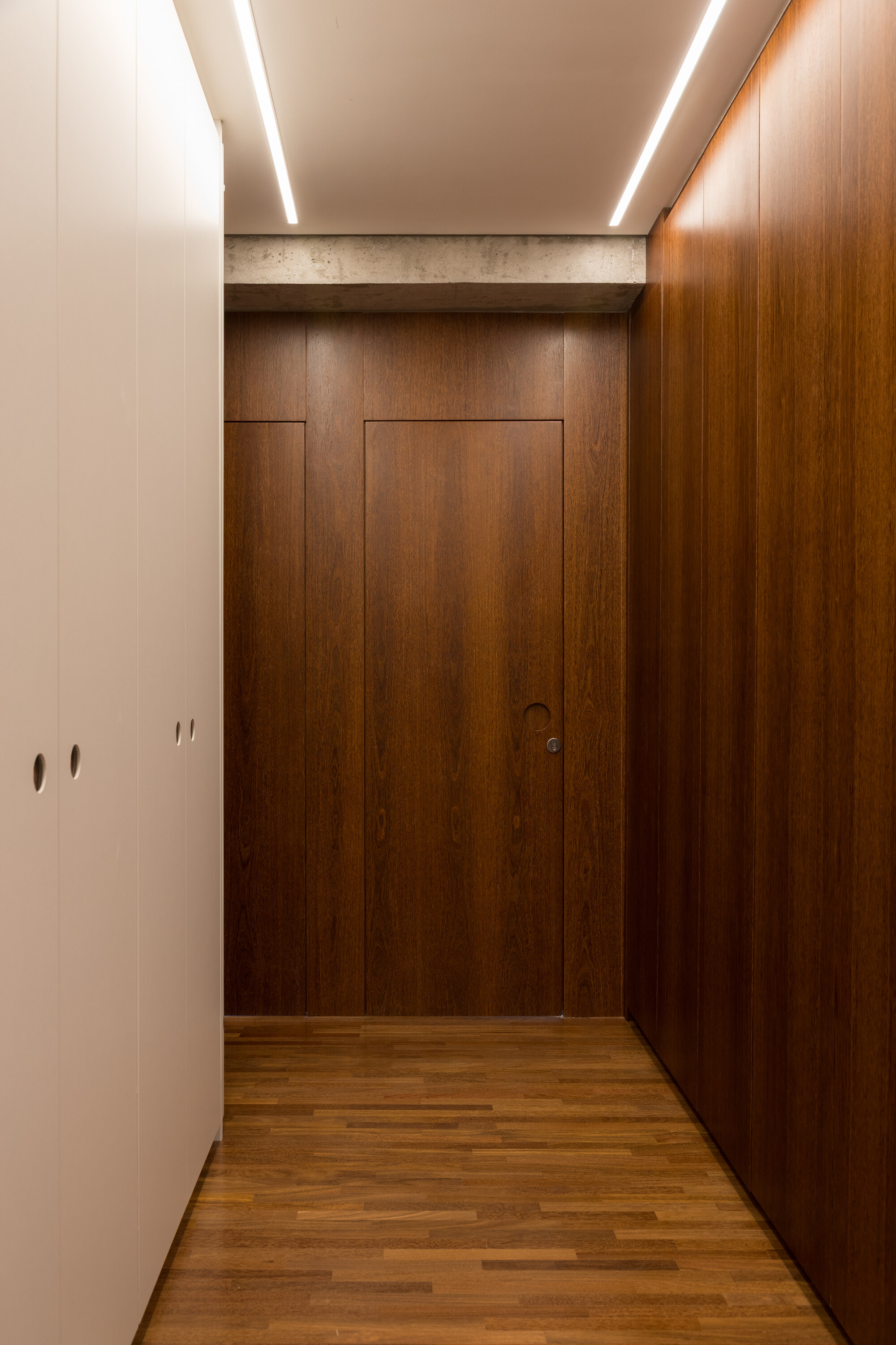 Narrow dressing corridor with white wardrobes, walnut wall panels, a flush door and linear ceiling lights above a parquet floor