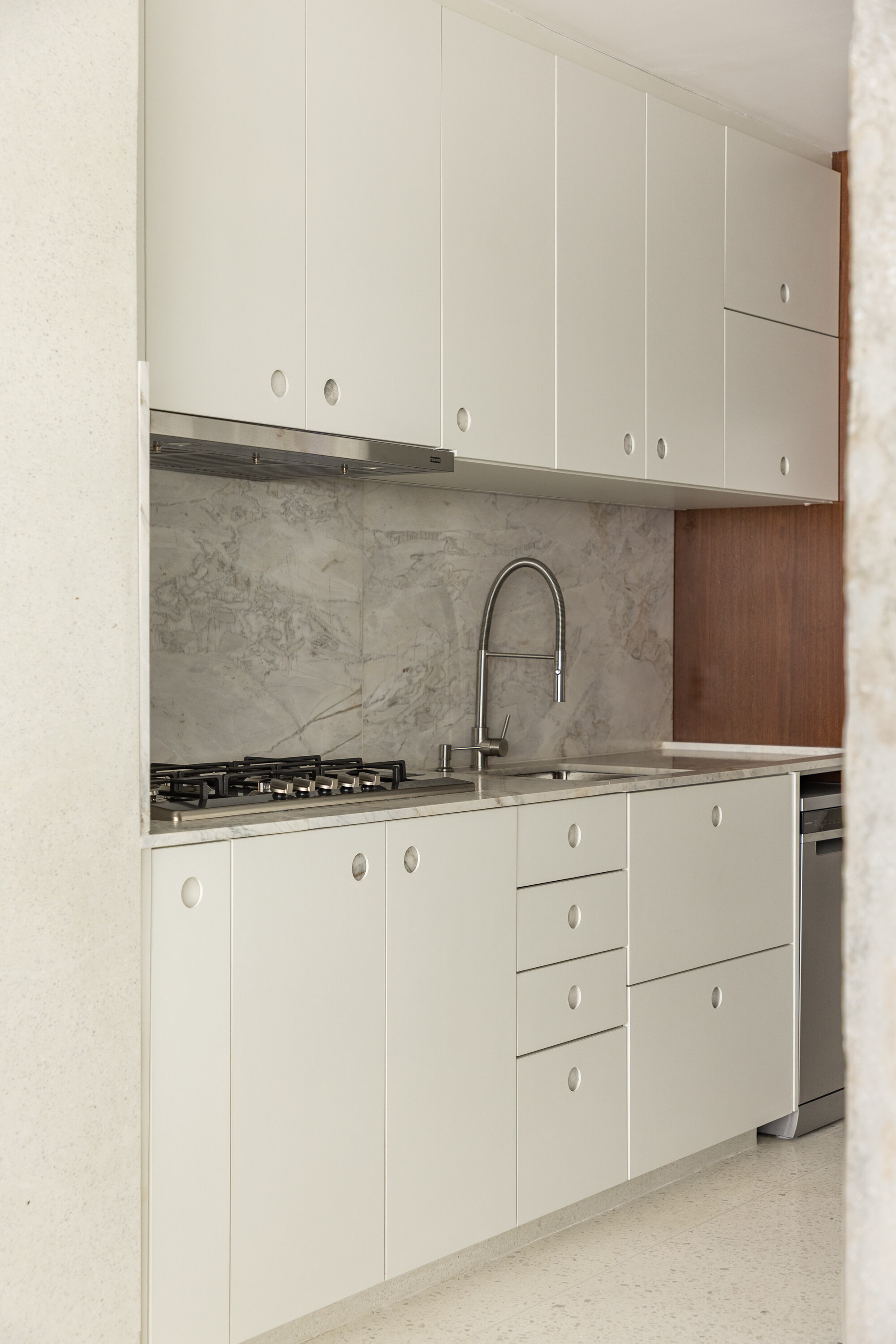 Compact kitchen run with white cabinets, round cut-out pulls, a gas hob and a grey marble backsplash behind a coiled tap
