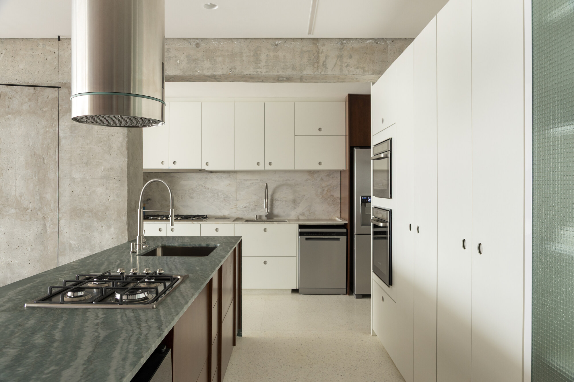 Kitchen with green stone island and cooktop, white cabinetry, steel hood and raw concrete wall in soft daylight