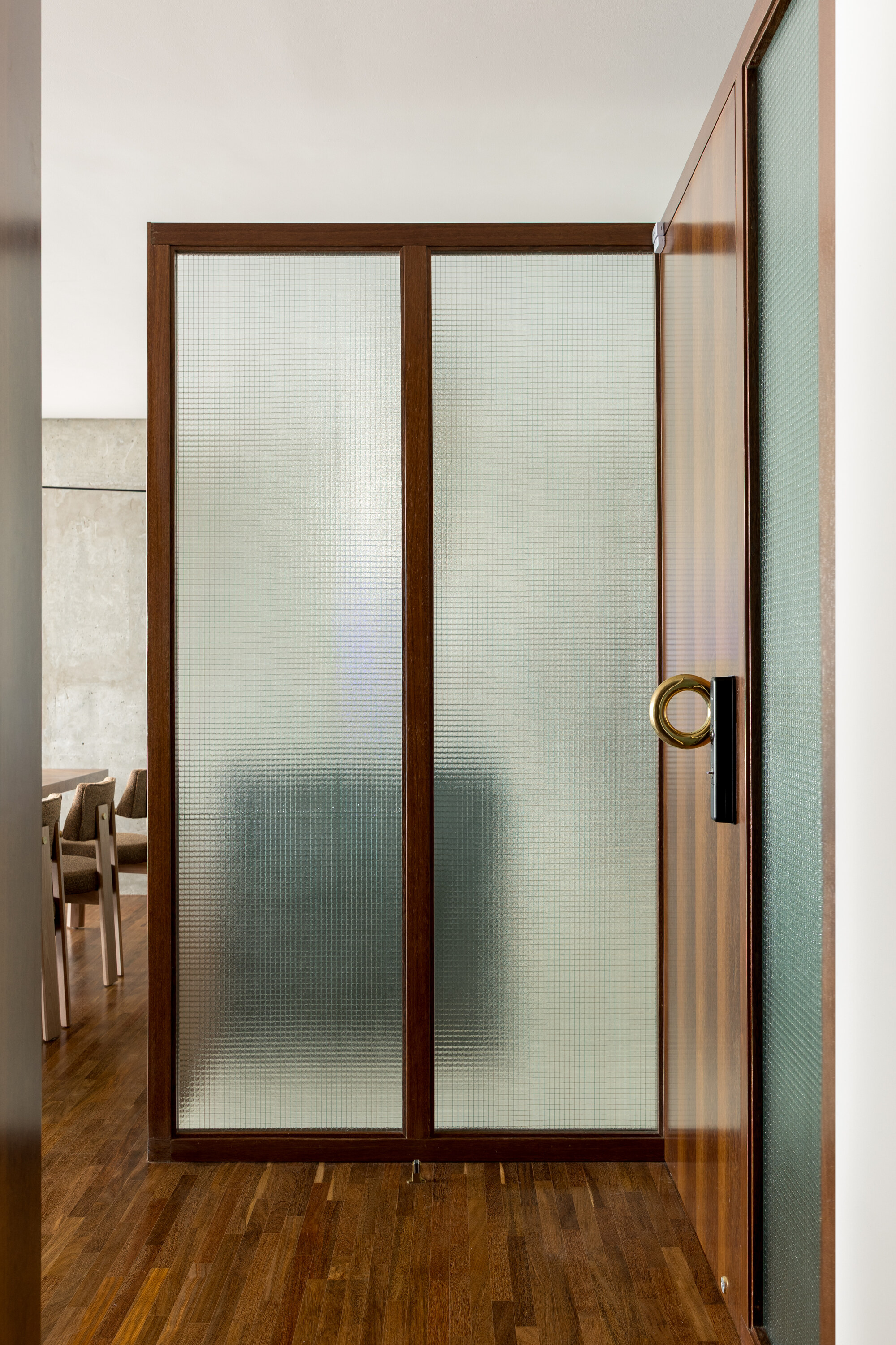 Wood-framed sliding partition with ribbed translucent glass and a brass ring pull beside a timber floor and dining chairs