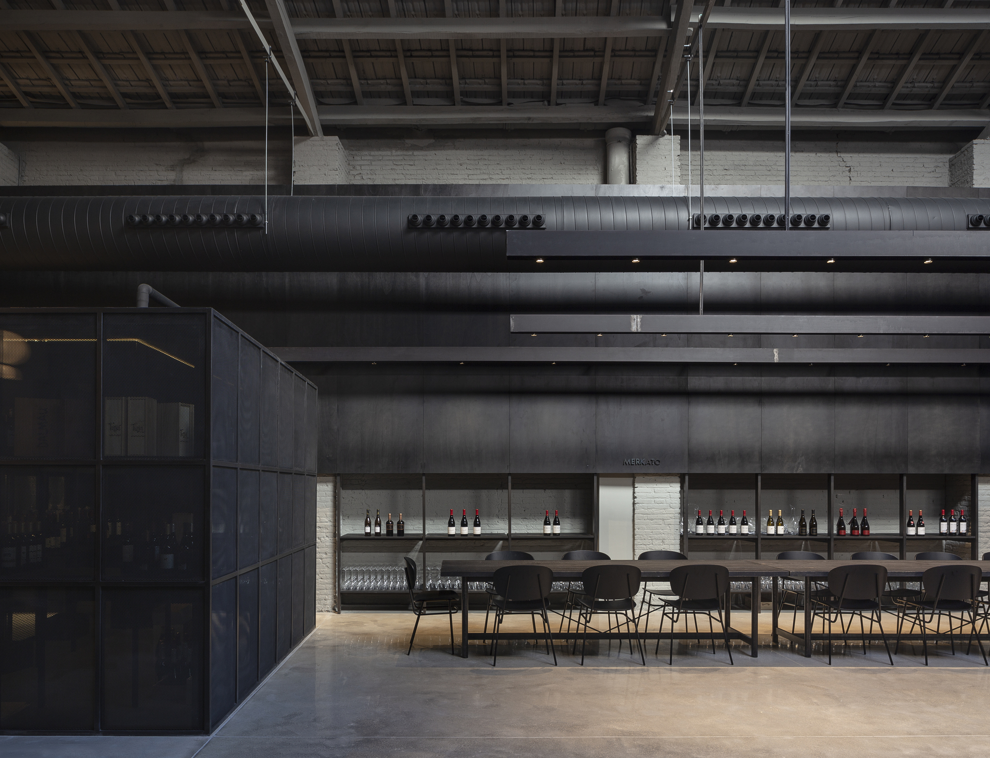 Long dining table and wine shelves beneath exposed timber roof and black ductwork, beside a dark mesh-clad cube