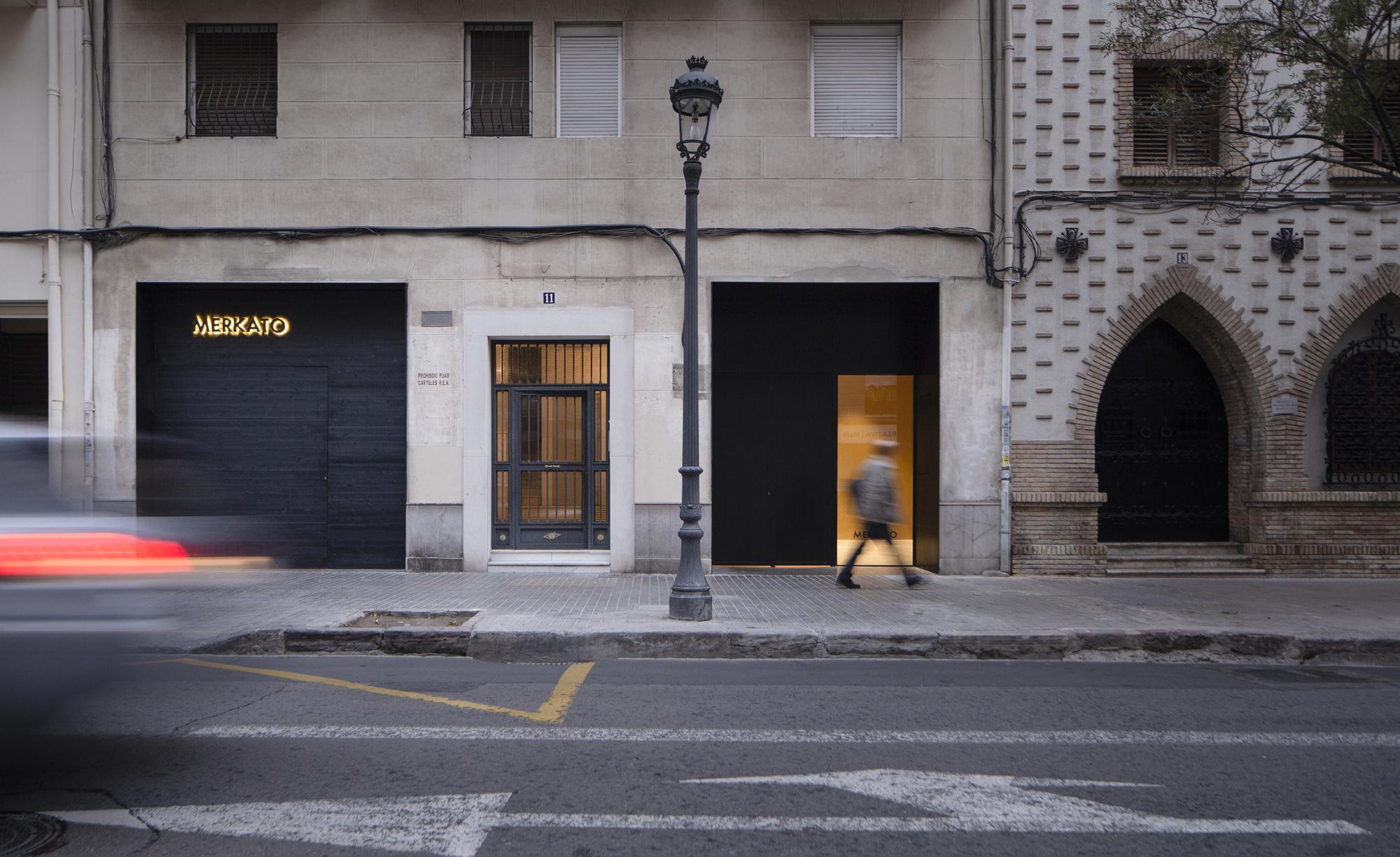 Street facade with black panel doors and a lit sign, a pedestrian blurred in motion and a car streaking past