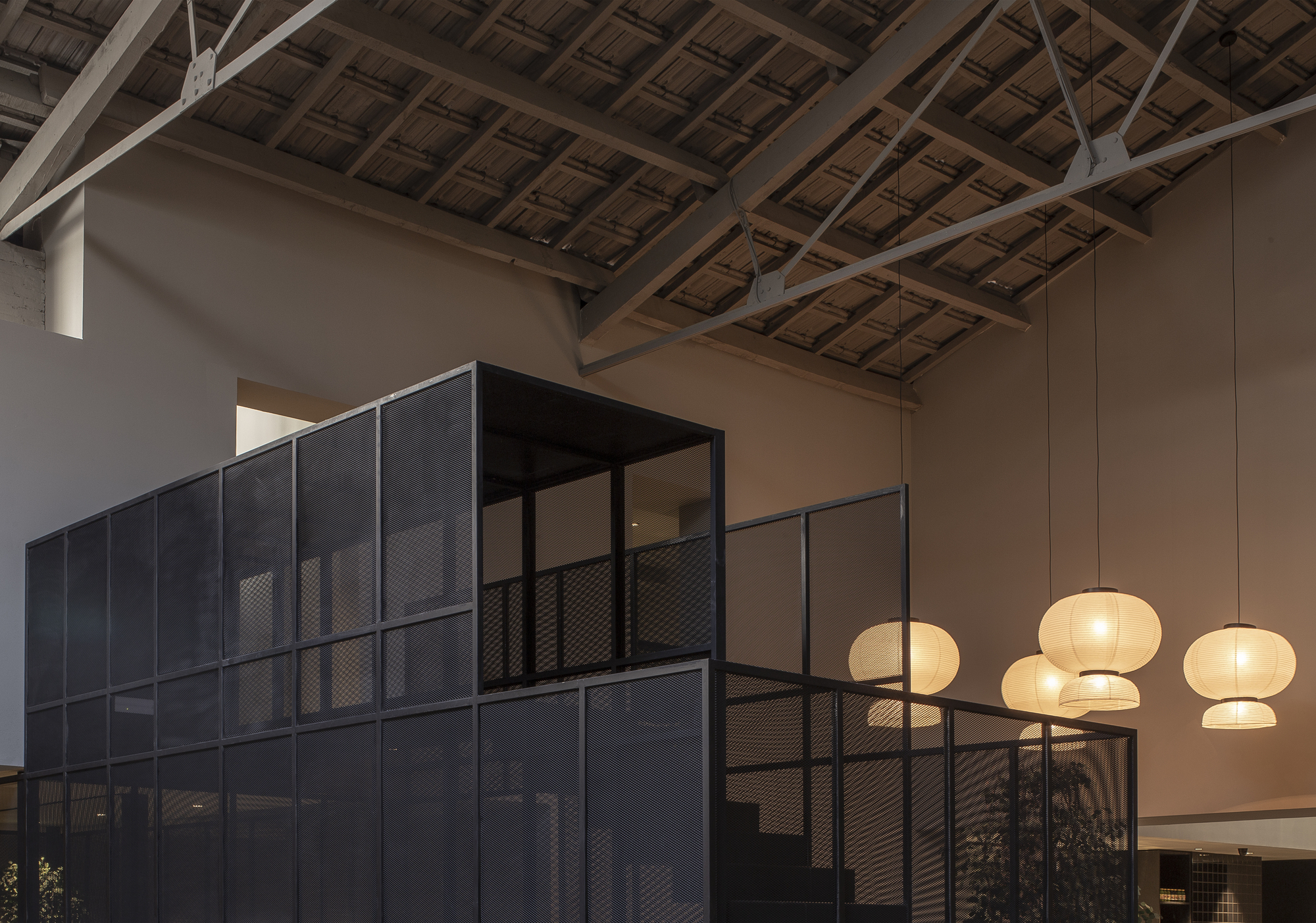 Black metal mesh volume with a glazed upper room under timber roof trusses, paper lantern pendants glowing at dusk