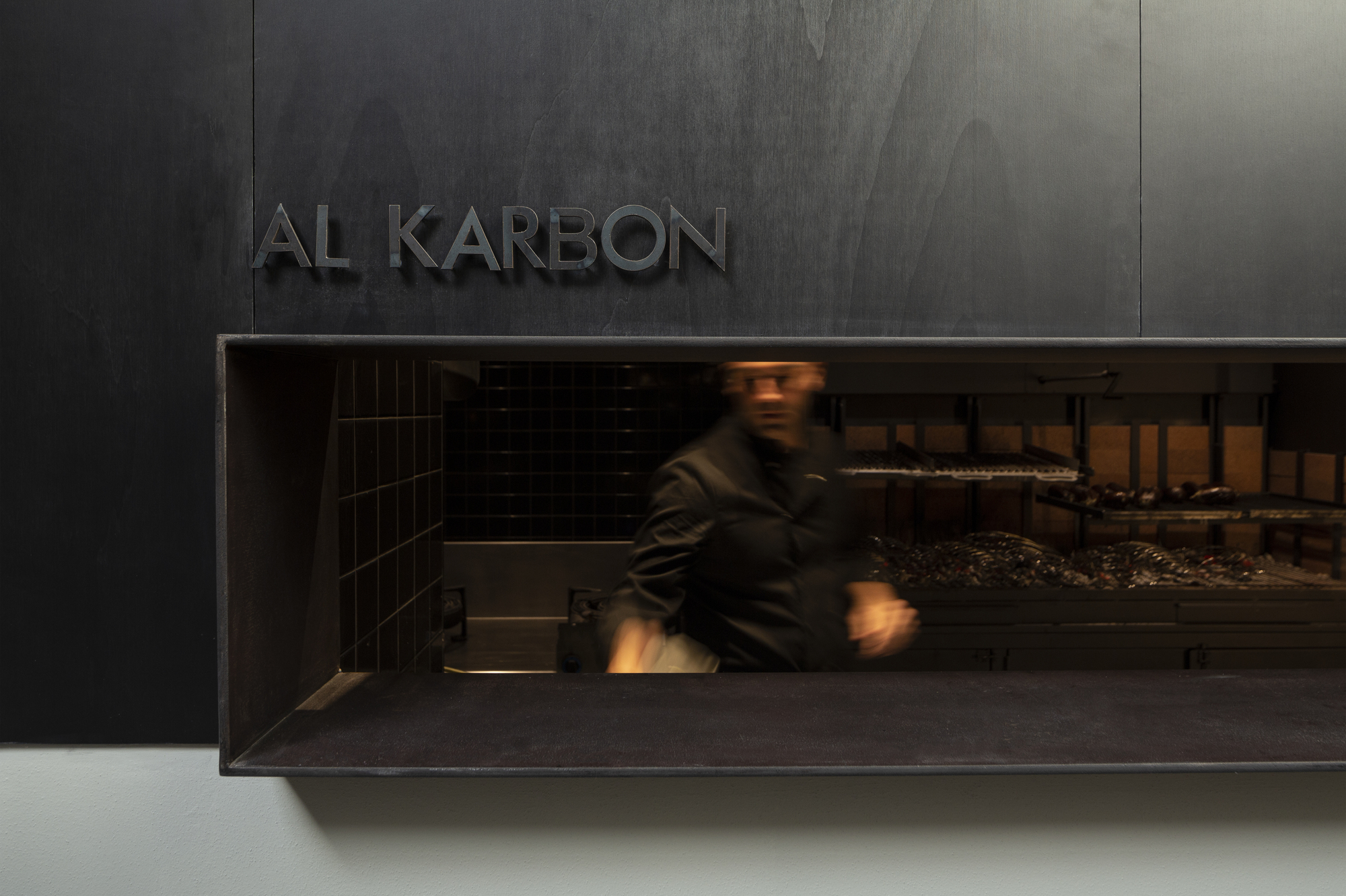 Serving hatch cut into a dark plywood wall, with a blurred cook moving in front of the charcoal grill