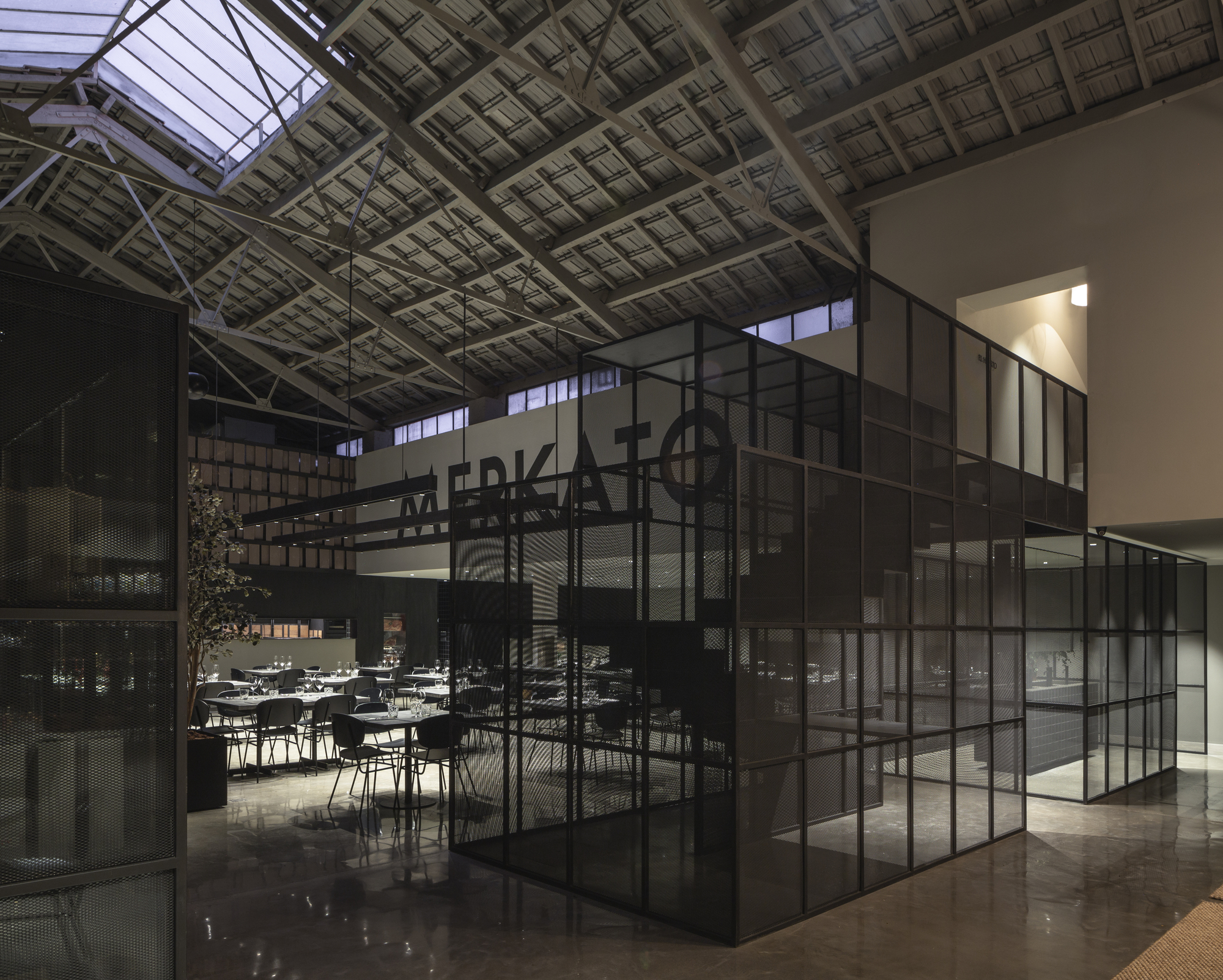 Glass and steel grid enclosure in a large hall with skylit timber trusses, dining tables glimpsed beyond in evening light