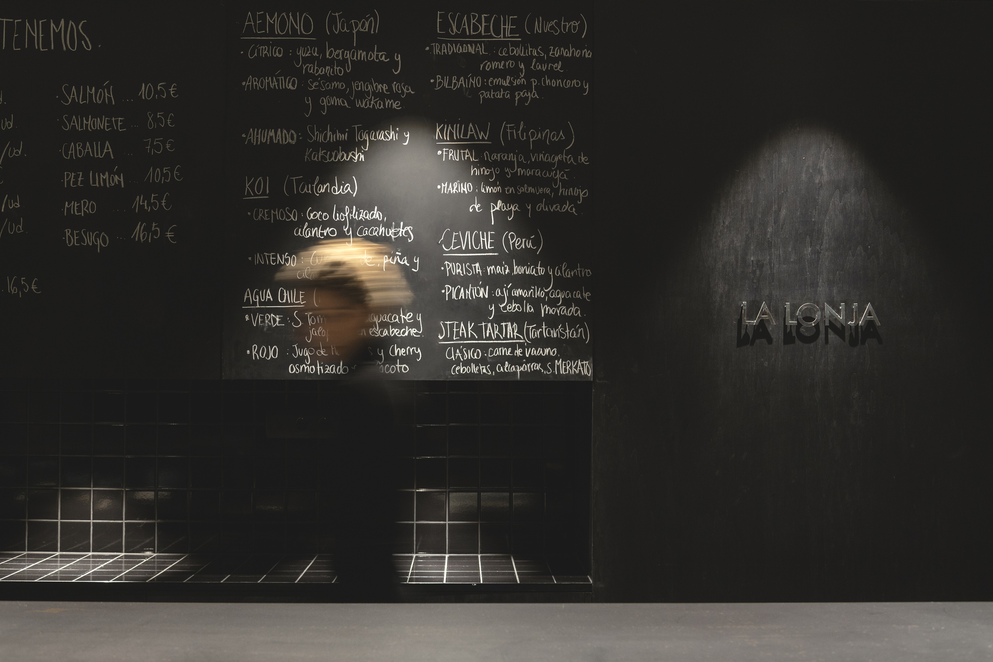 Chalkboard menu above black tiled counter, a passing figure blurred in a spotlight beside a metal wall sign