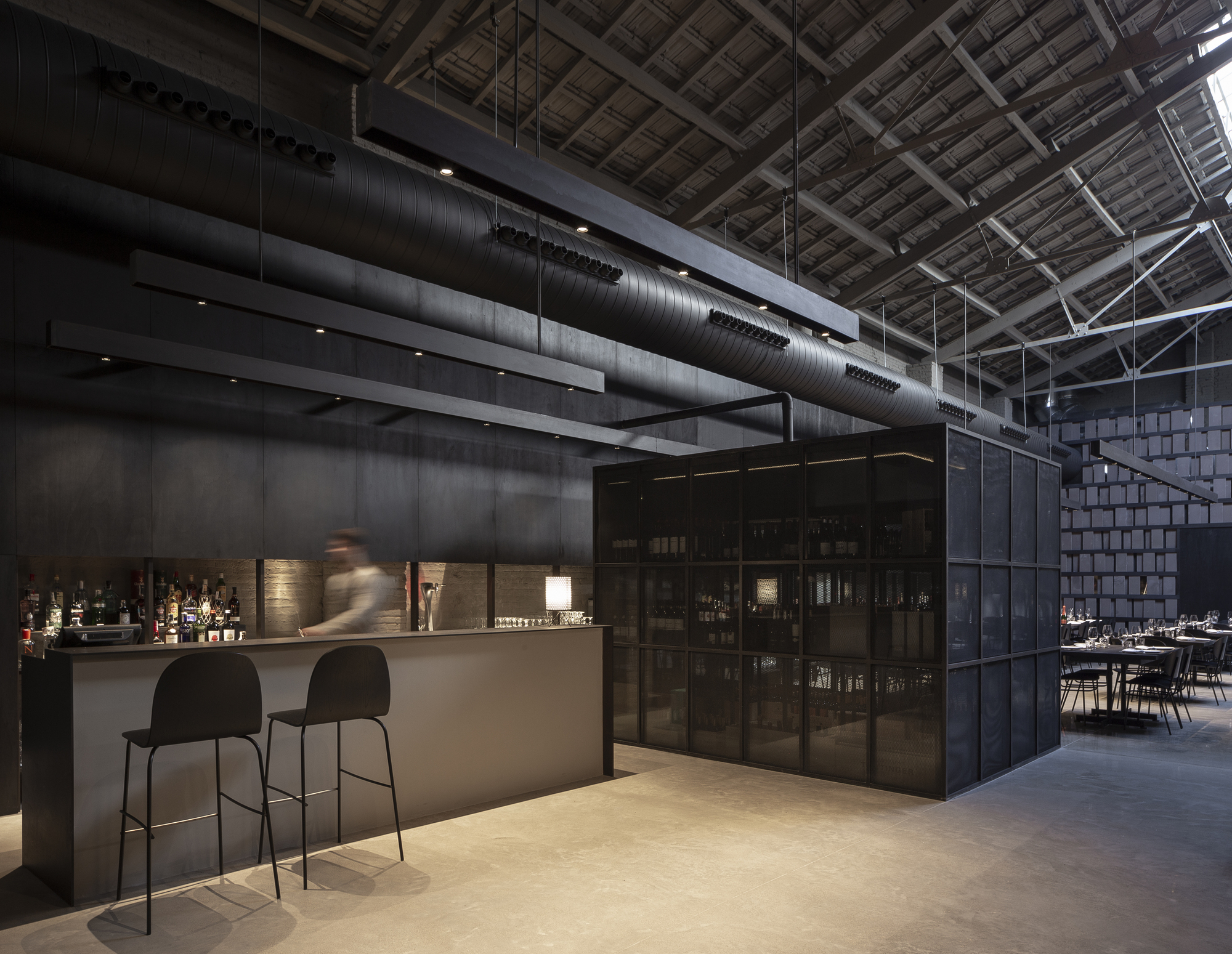 Black bar counter with two stools beside a mesh-clad wine cube, under exposed roof trusses and a blurred bartender