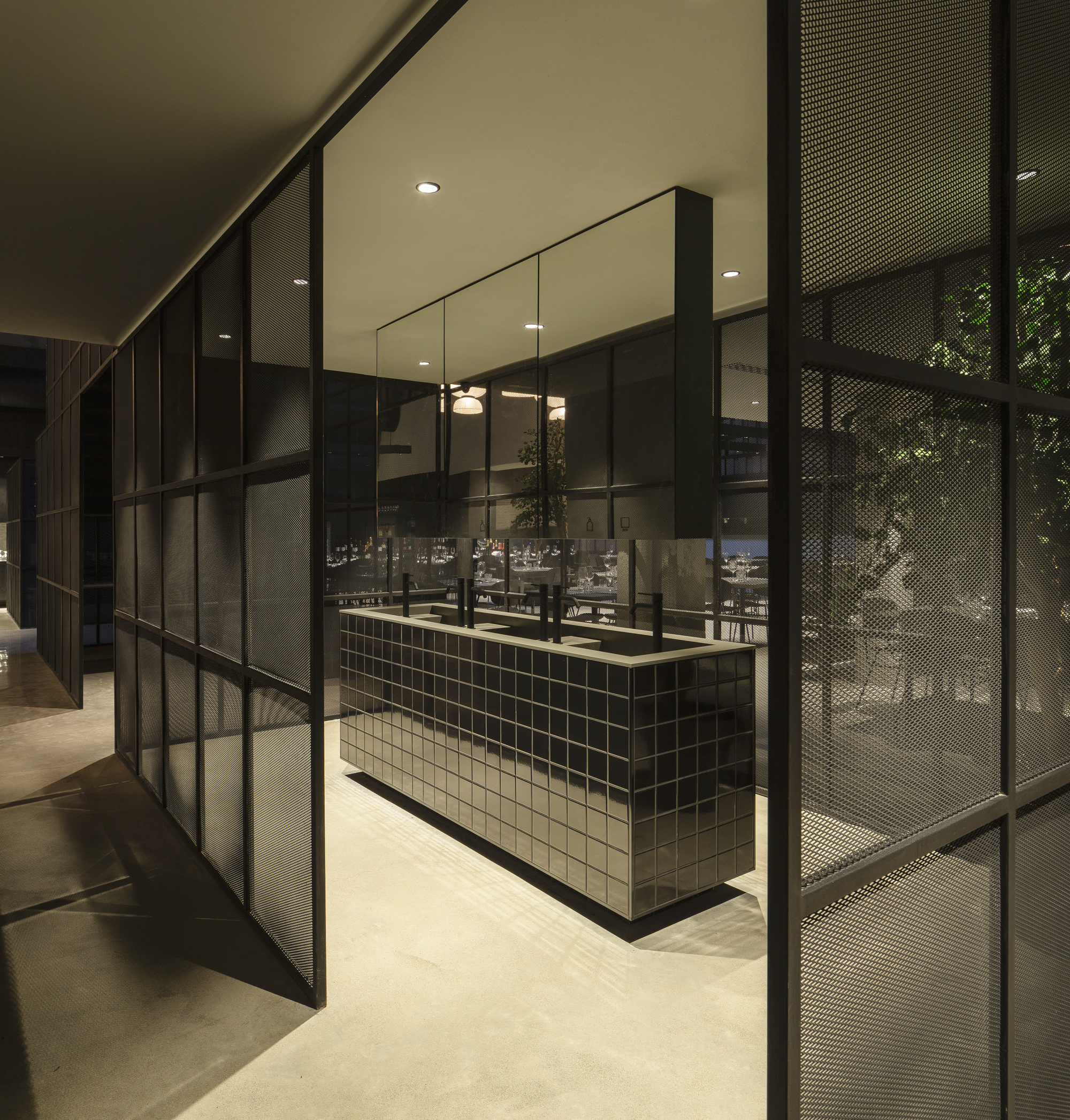 Mirror-tiled counter seen between steel-framed mesh partitions, with dining tables and foliage glowing in the evening dimness