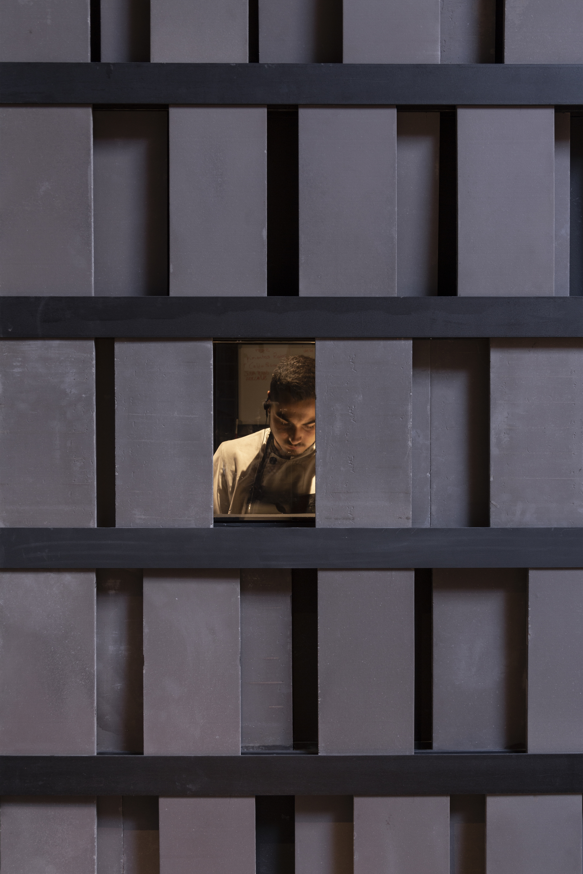 Close-up of dark timber slat screen with a small opening framing a cook working in the kitchen beyond