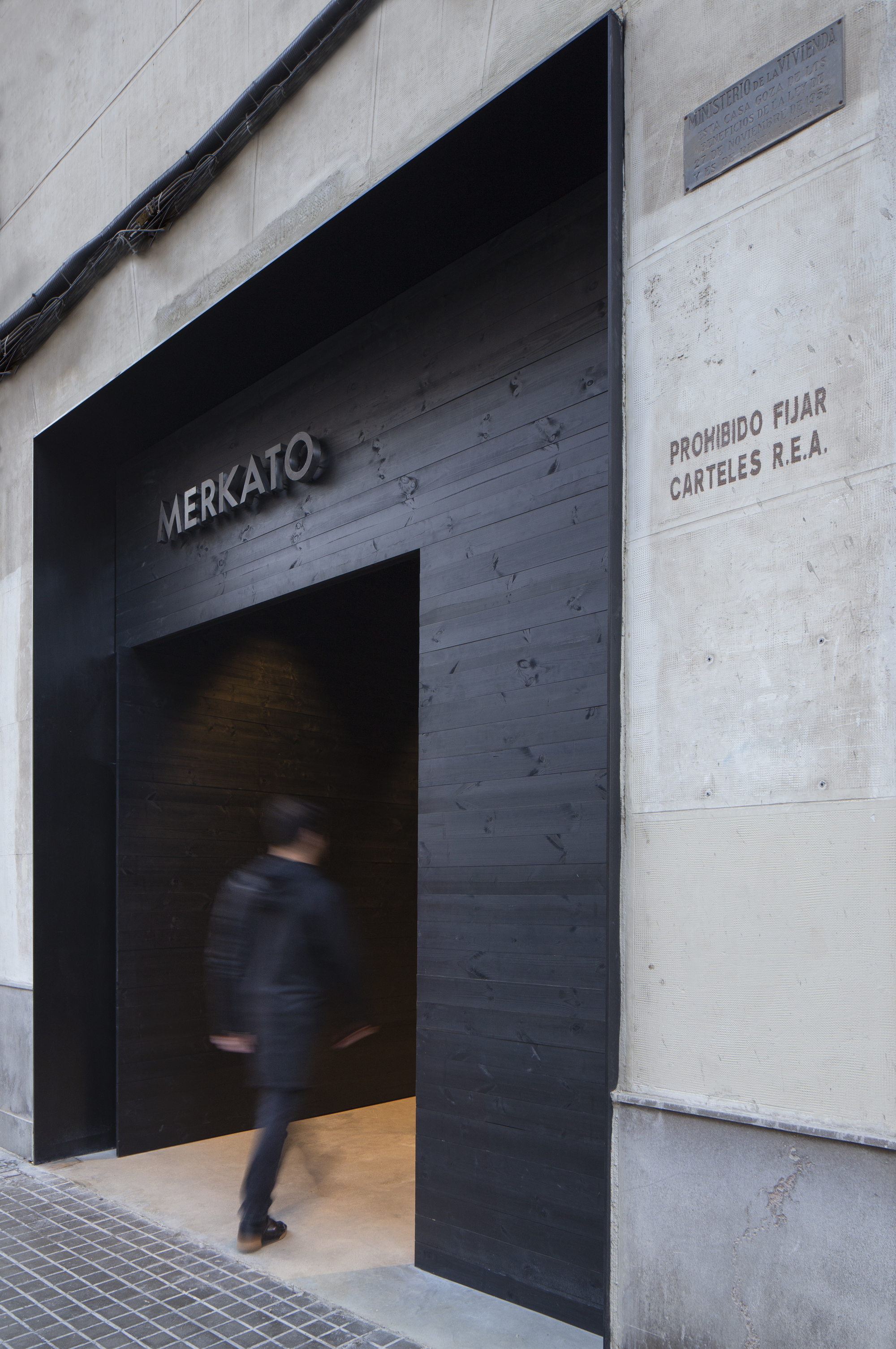 Entrance clad in charred black timber boards with a hanging sign, a pedestrian blurred in motion on the pavement