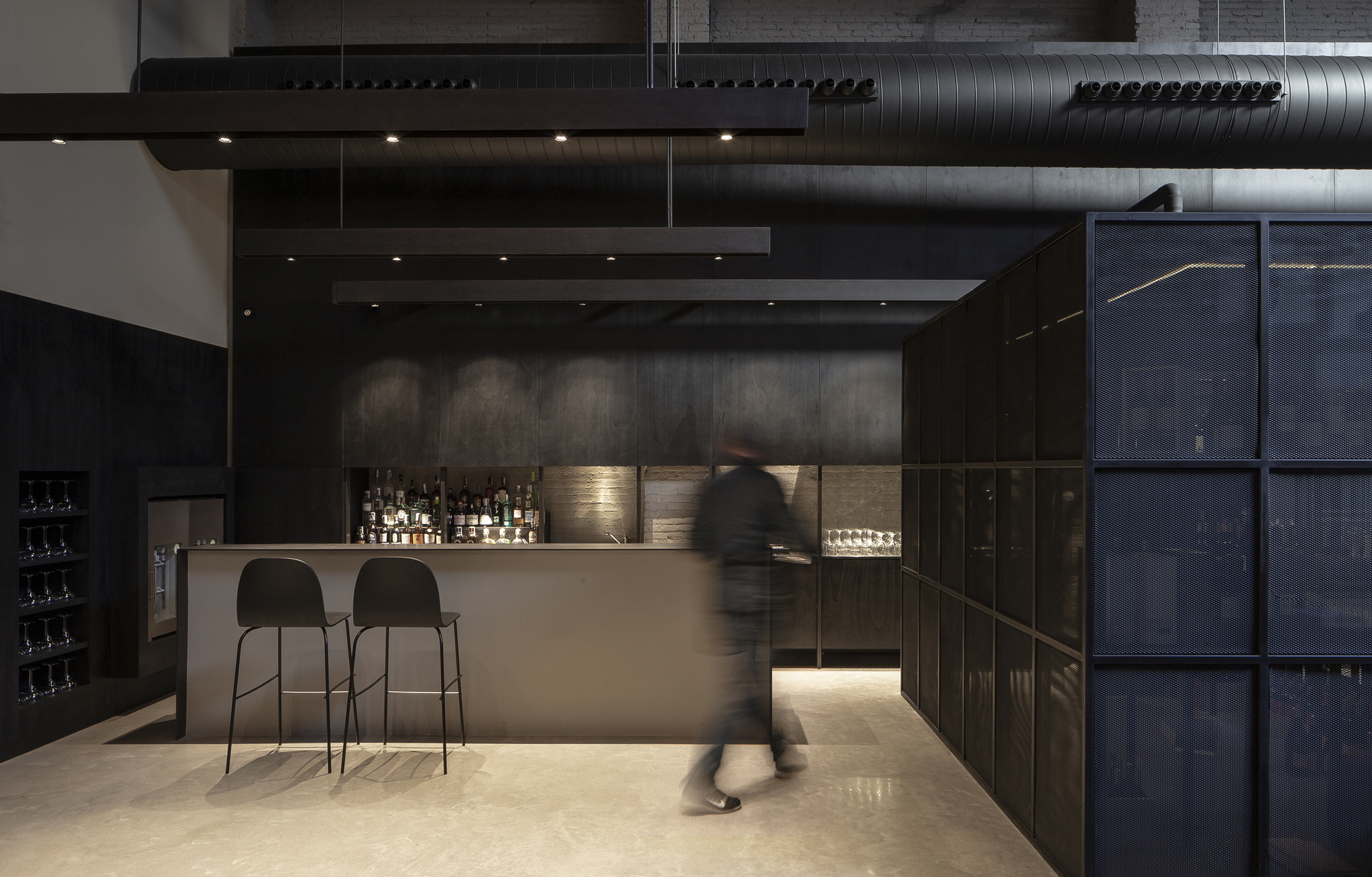 Bar counter with two black stools beside a mesh-panelled steel volume, a blurred staff member walking past under linear pendant lights