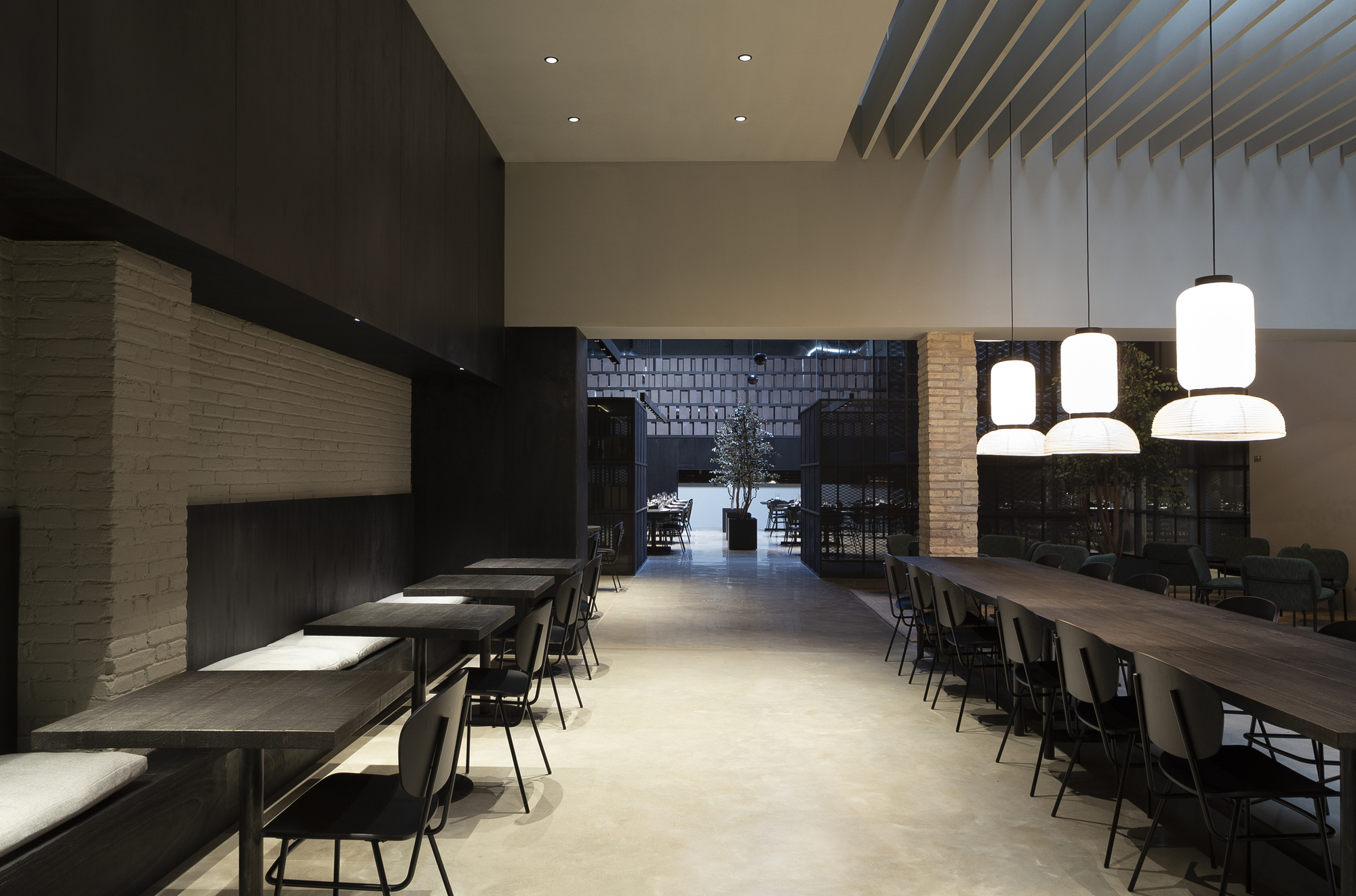 Dining hall with banquette seating along a painted brick wall and paper lantern pendants hanging over a long timber table