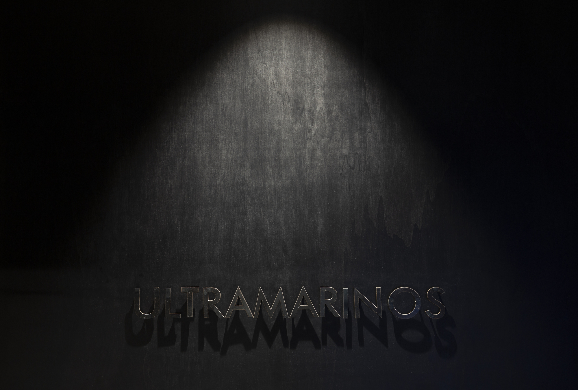 Metal letter signage on a dark textured wall, lit by a single downward spotlight that casts a shadow