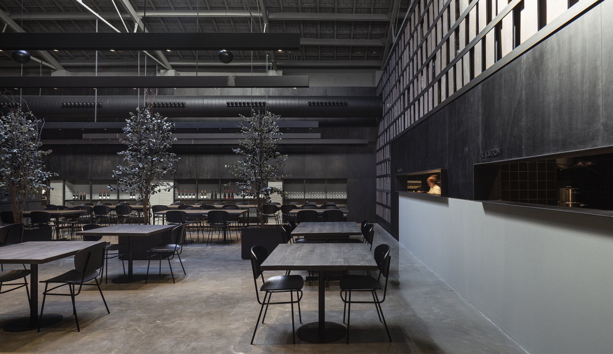 Dining room of square tables and black chairs with potted trees, facing an open kitchen window in a dark wall