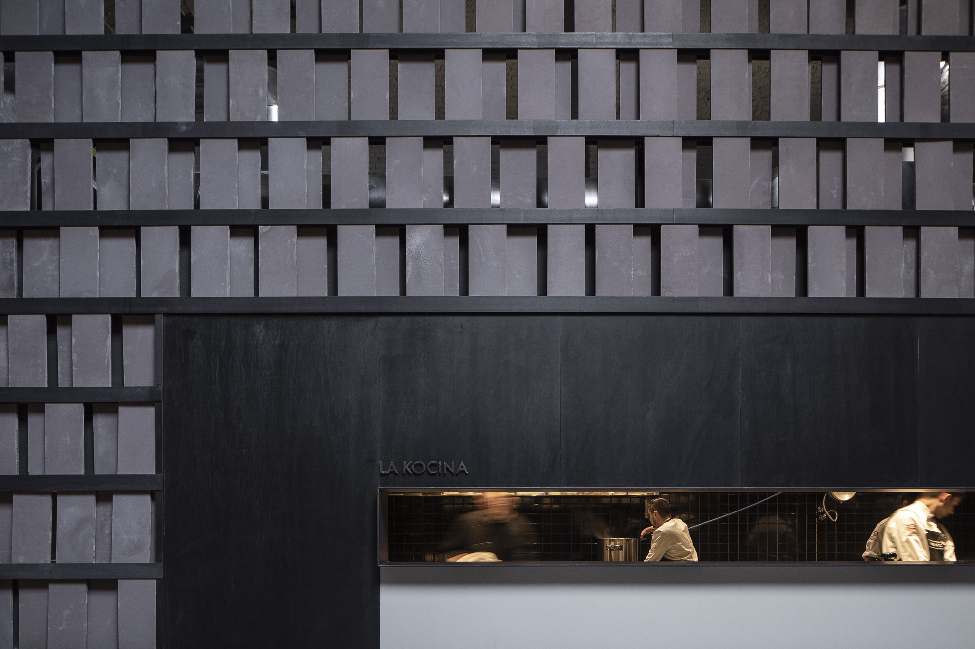 Close-up of the slatted grey brick screen above the open kitchen window, with cooks in white jackets visible inside