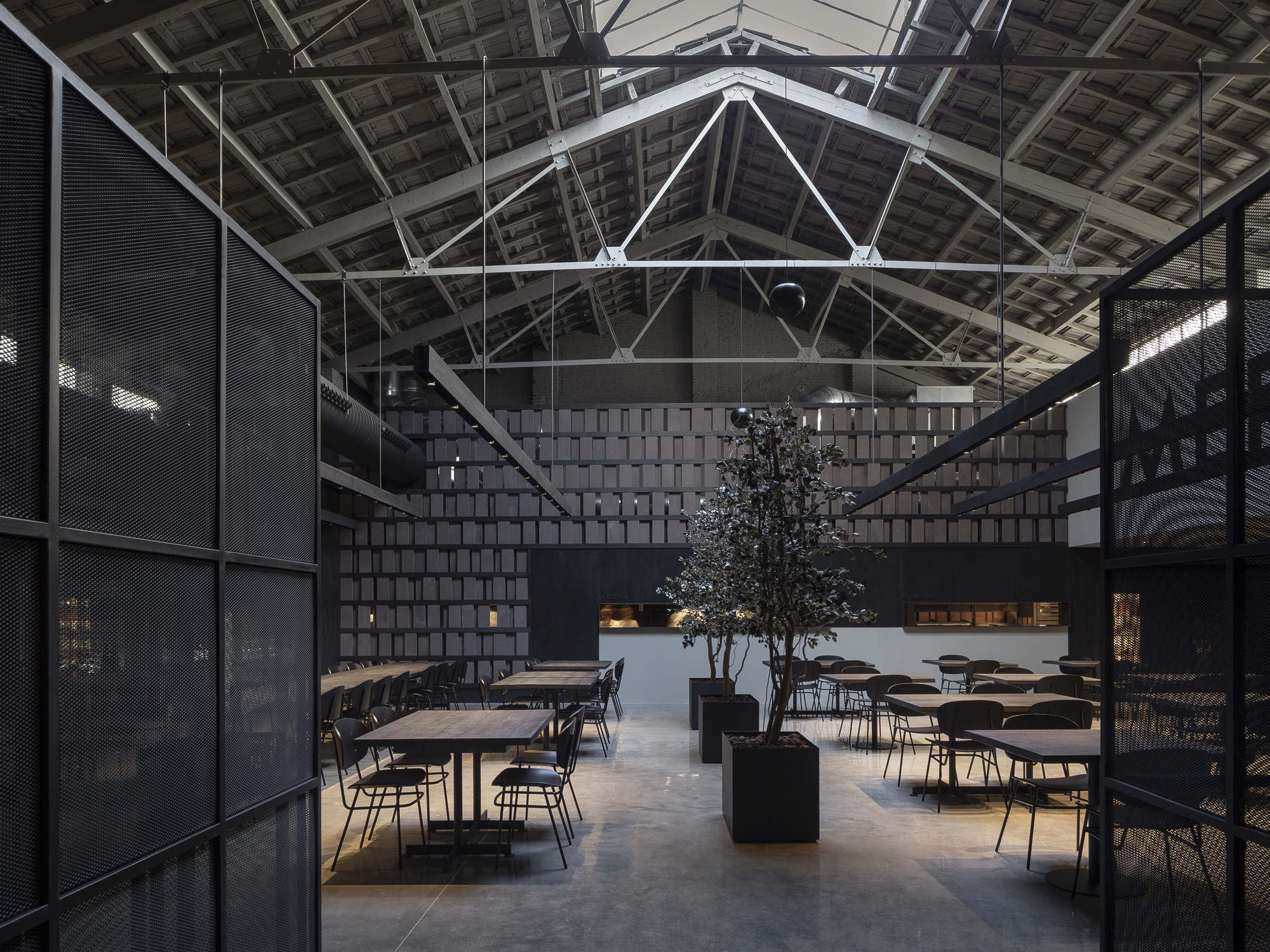Restaurant under exposed steel roof trusses, with potted olive trees and tables seen between black metal mesh screens in soft daylight