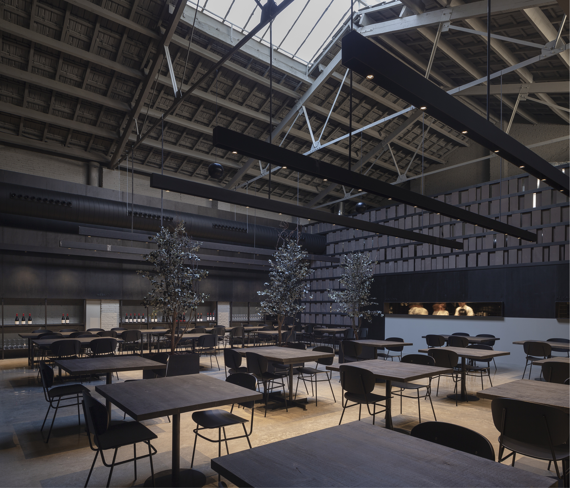 Restaurant hall with dark wood tables, potted trees and suspended black light bars beneath exposed steel roof trusses and a skylight