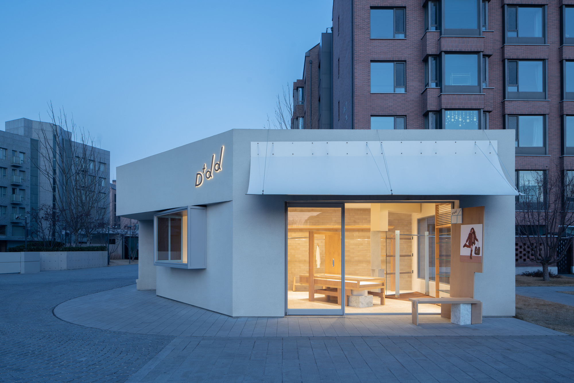 Small white-rendered pavilion with a canopy awning and glowing storefront at dusk, beside a brick apartment block