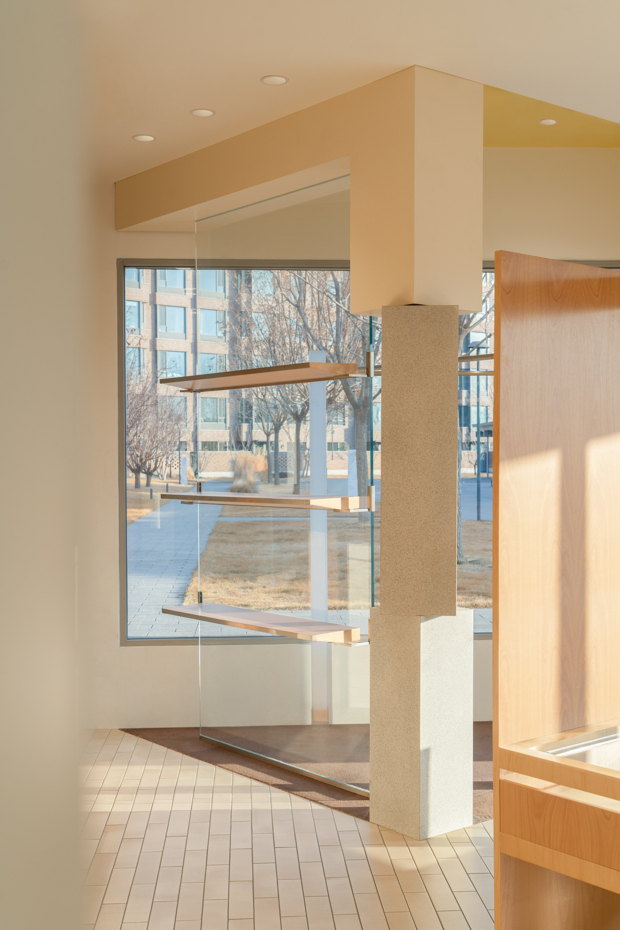 Speckled stone column below a boxed beam beside a glazed corner with wooden shelves, bare trees outside in winter light
