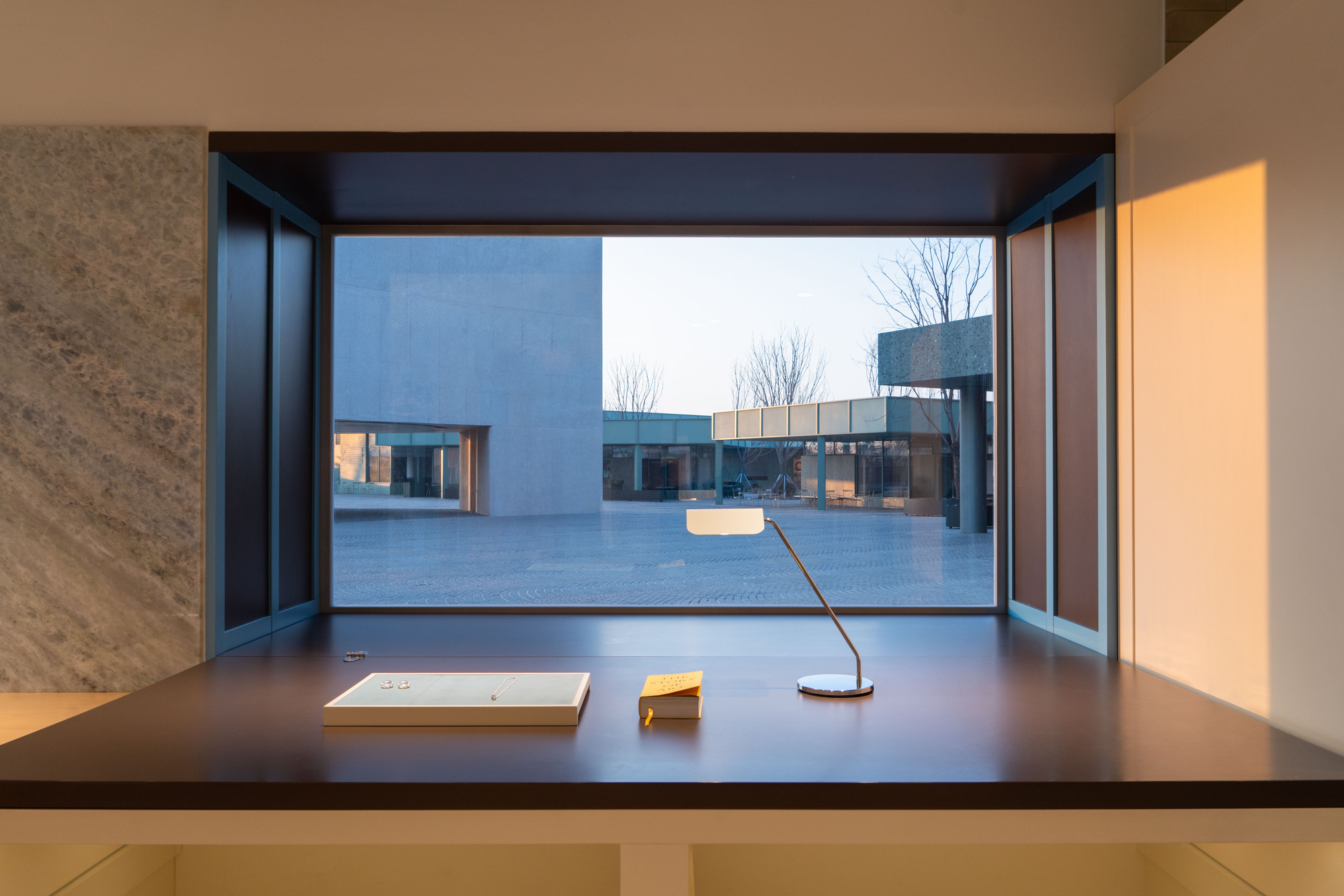 Deep window seat desk framing a paved courtyard and concrete volume, with a desk lamp and books in low evening sun