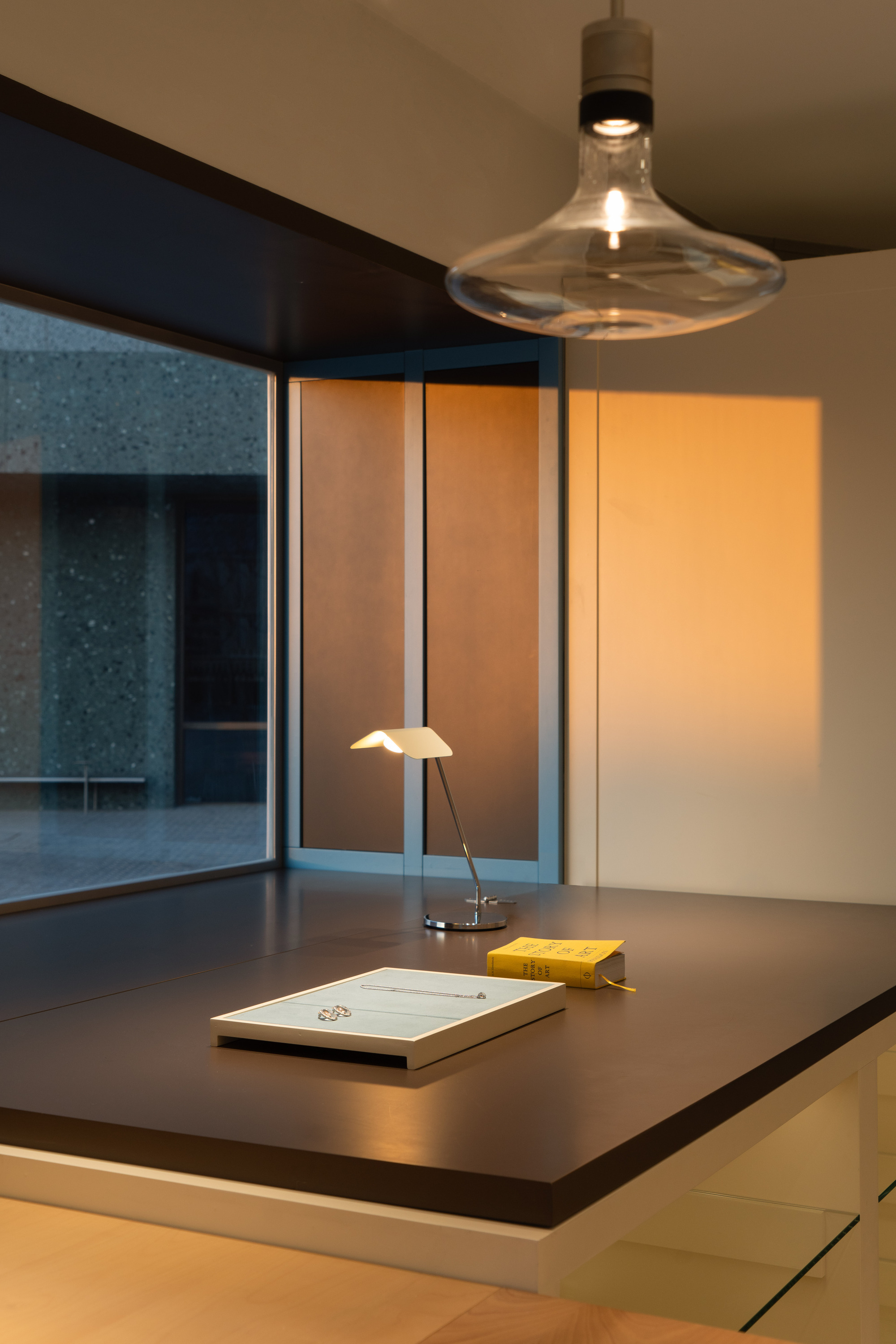 Dark desk surface with lamp, yellow book and glass pendant, warm sunset light falling on the white wall beside the window
