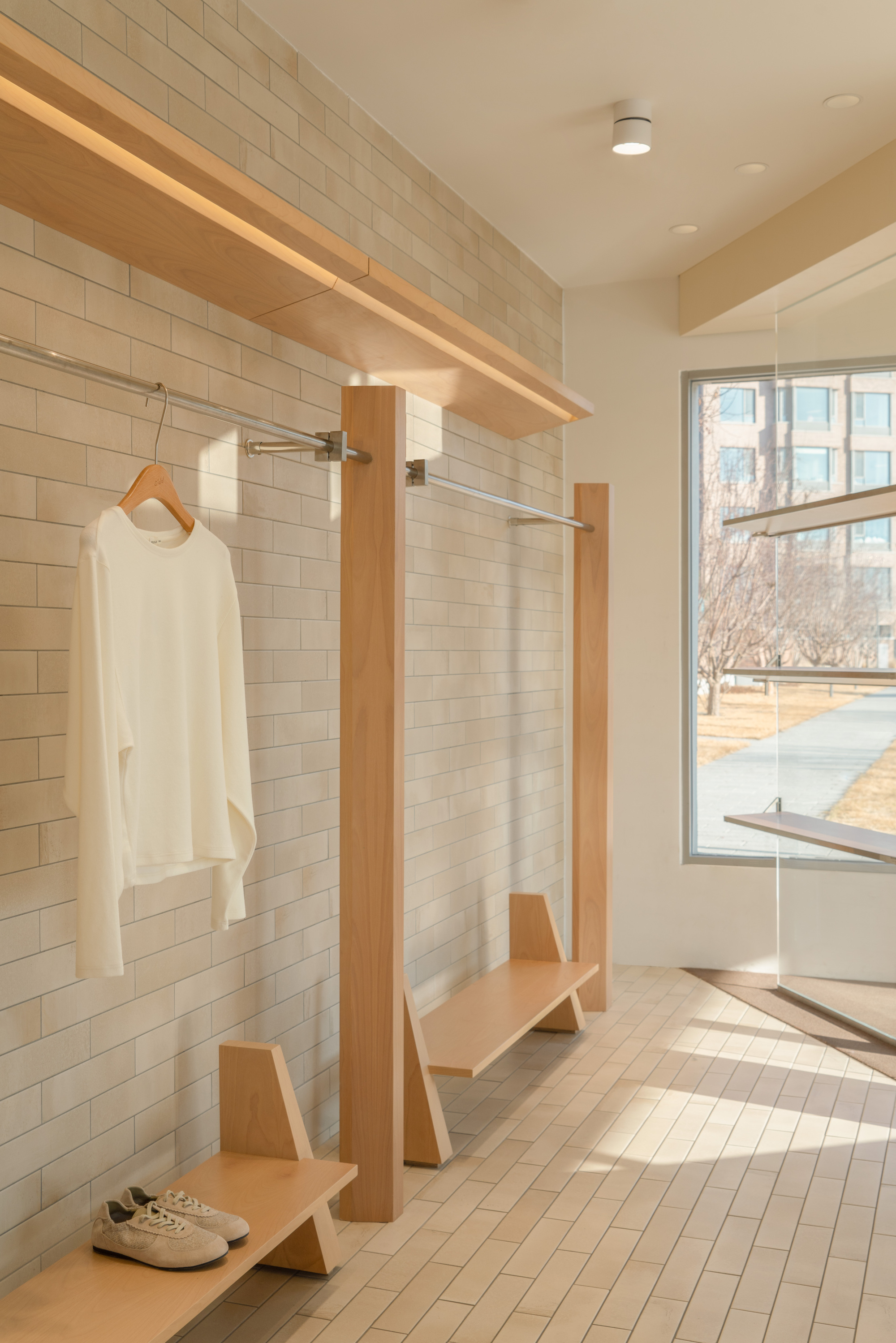 Garment rail with timber posts and built-in benches against a pale brick wall, sunlight striping the tiled floor