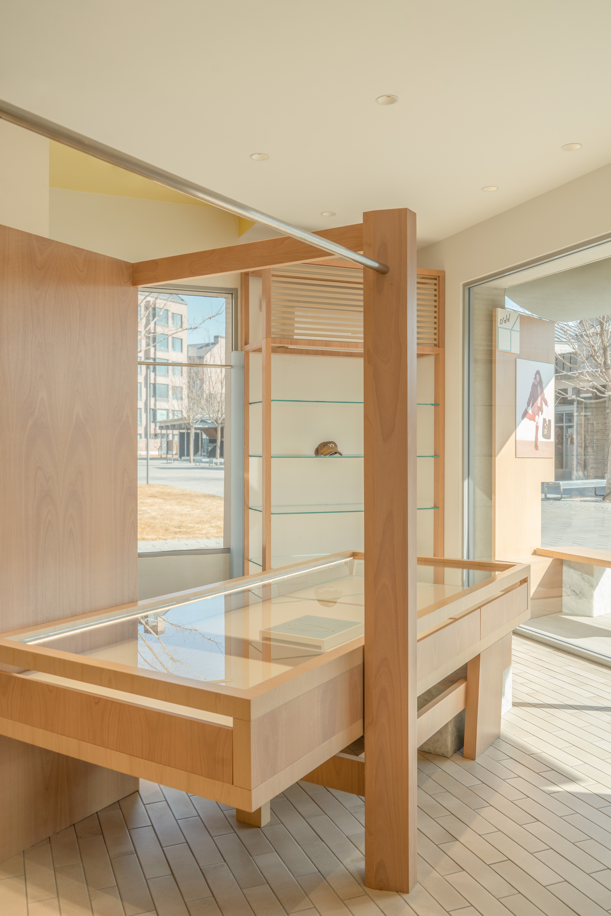 Timber display table with glass top beside a wood post and glass-shelved cabinet, with winter sunlight falling across a pale plank floor