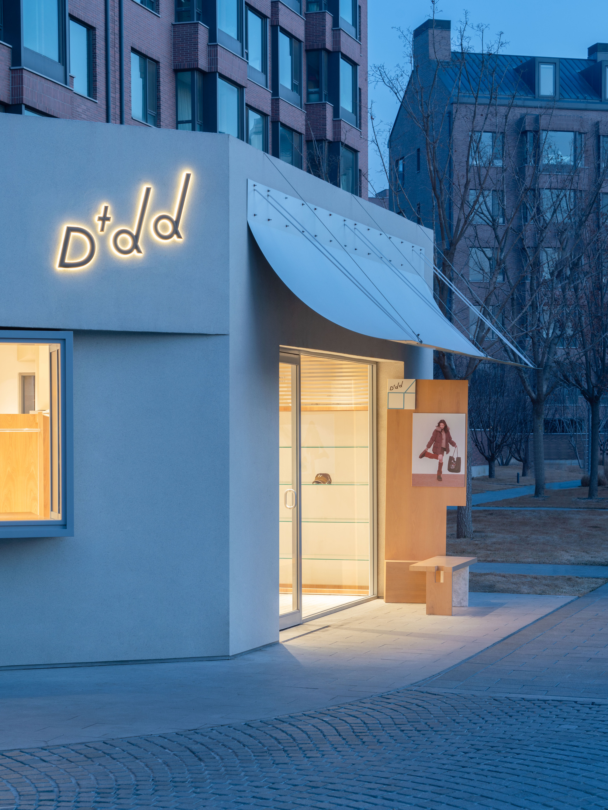 Shopfront corner with illuminated script sign and curved steel canopy over a glazed door, seen at dusk beside bare trees