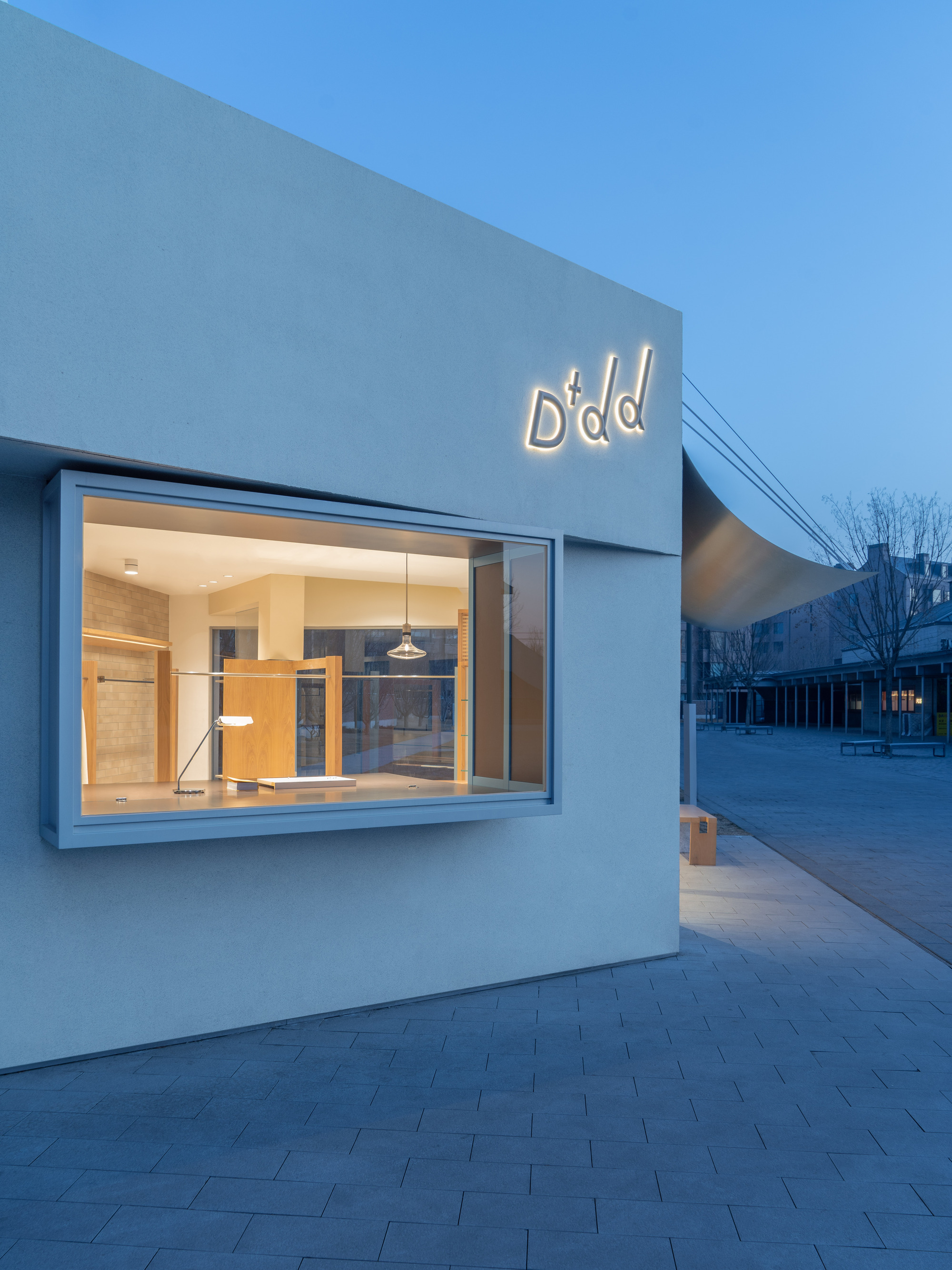 Projecting box window in a pale rendered wall, glowing with warm interior light under a deep blue dusk sky