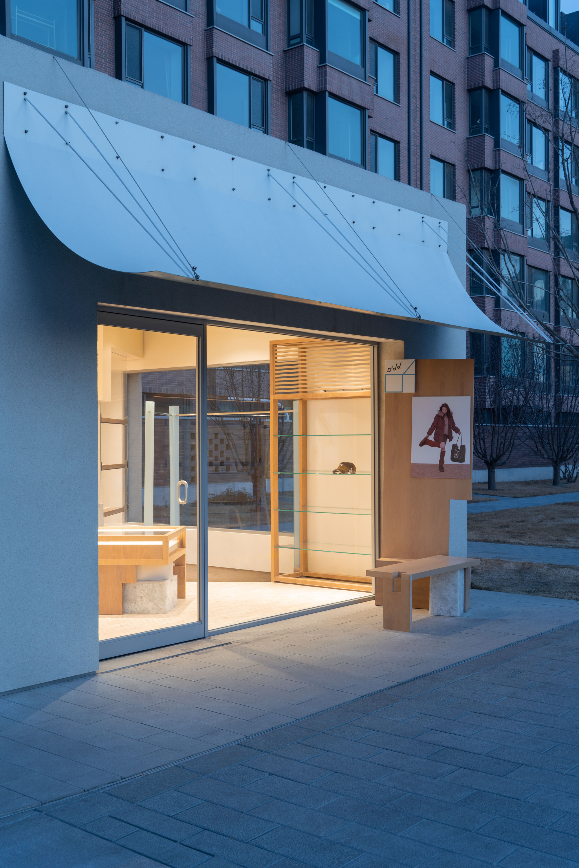 Curved metal canopy with tension rods above the glazed entrance and a wood and stone bench, lit at twilight