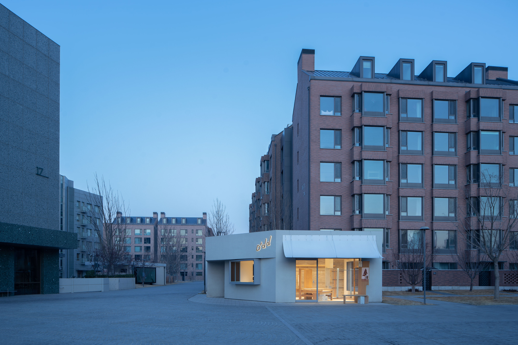 Small pale pavilion with glowing glazed front standing in an open paved square between brick apartment blocks at blue hour