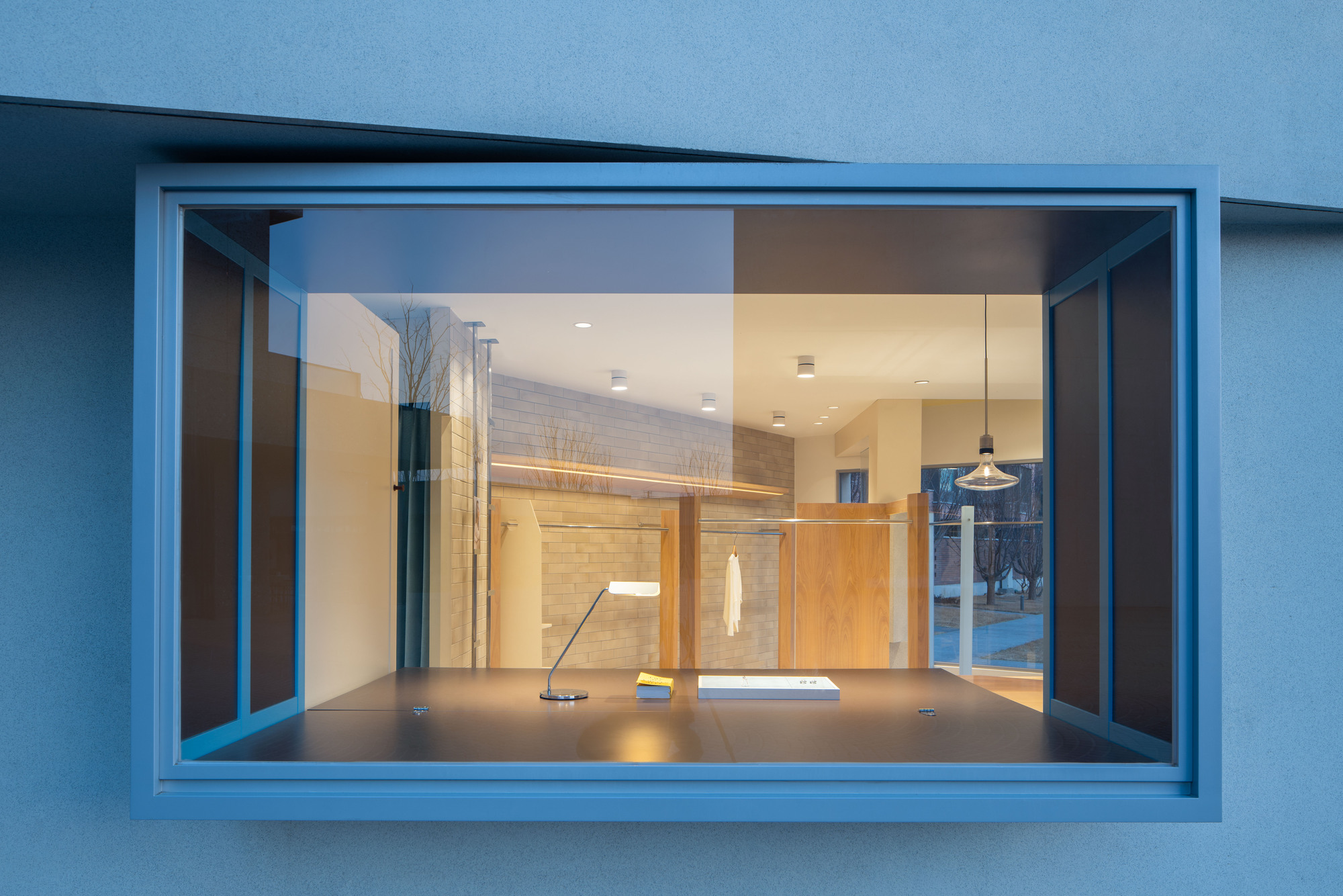 Close view through the framed box window to a dark display counter, desk lamp and pendant light at dusk