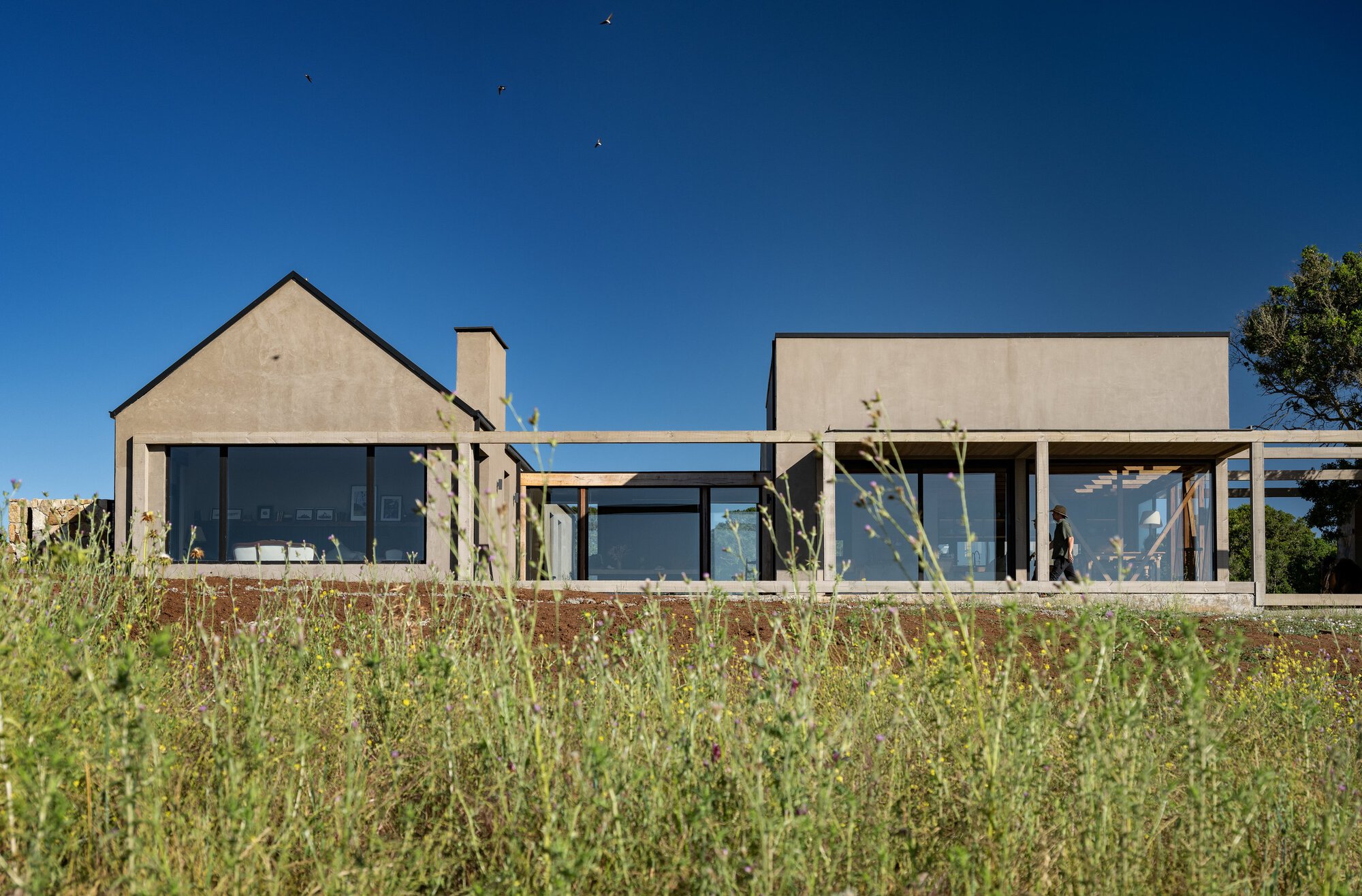 Low gabled stucco house behind a timber pergola frame, seen across tall wild grasses with a person walking under deep blue sky